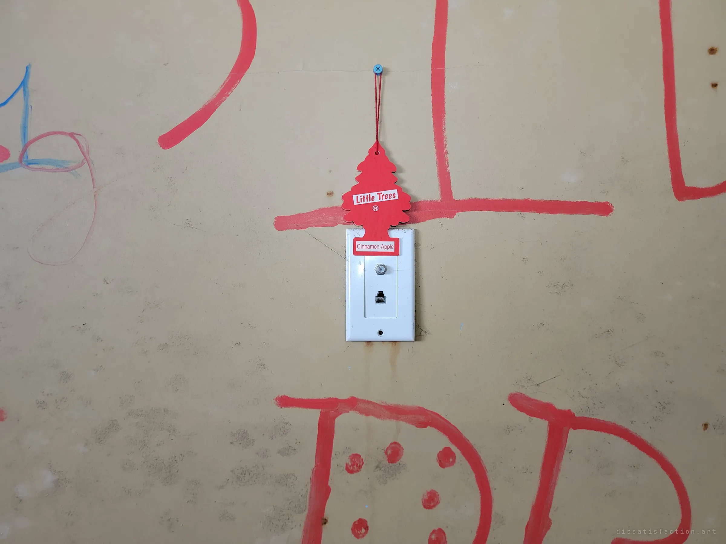 A red tree-shaped air freshener hung on a screw above a cable and phone wall plate, framed by big red painted letters across the bare wall.