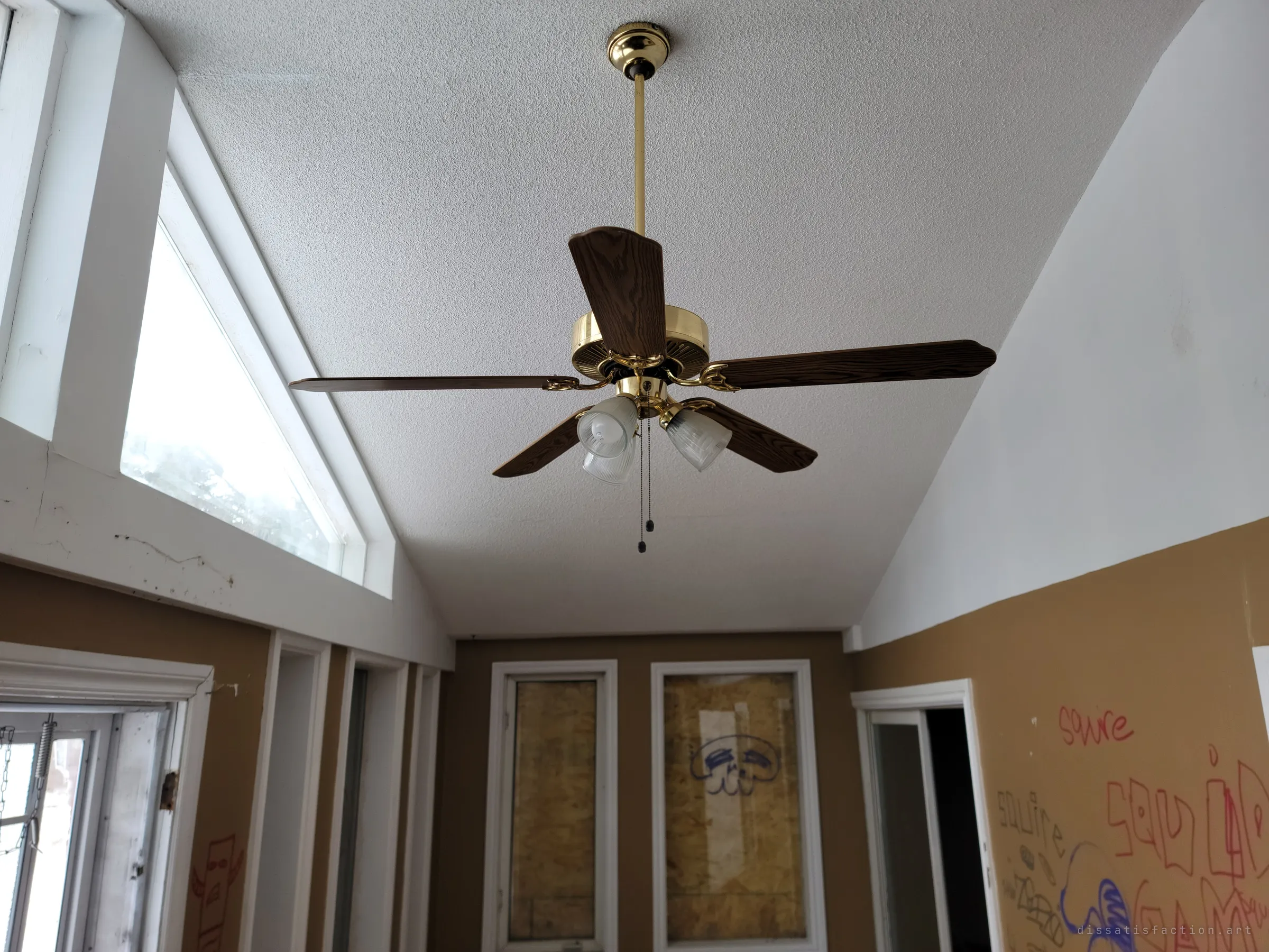 A brass ceiling fan still hangs in a vaulted sunroom whose tall windows are boarded, the walls below scrawled with tags.