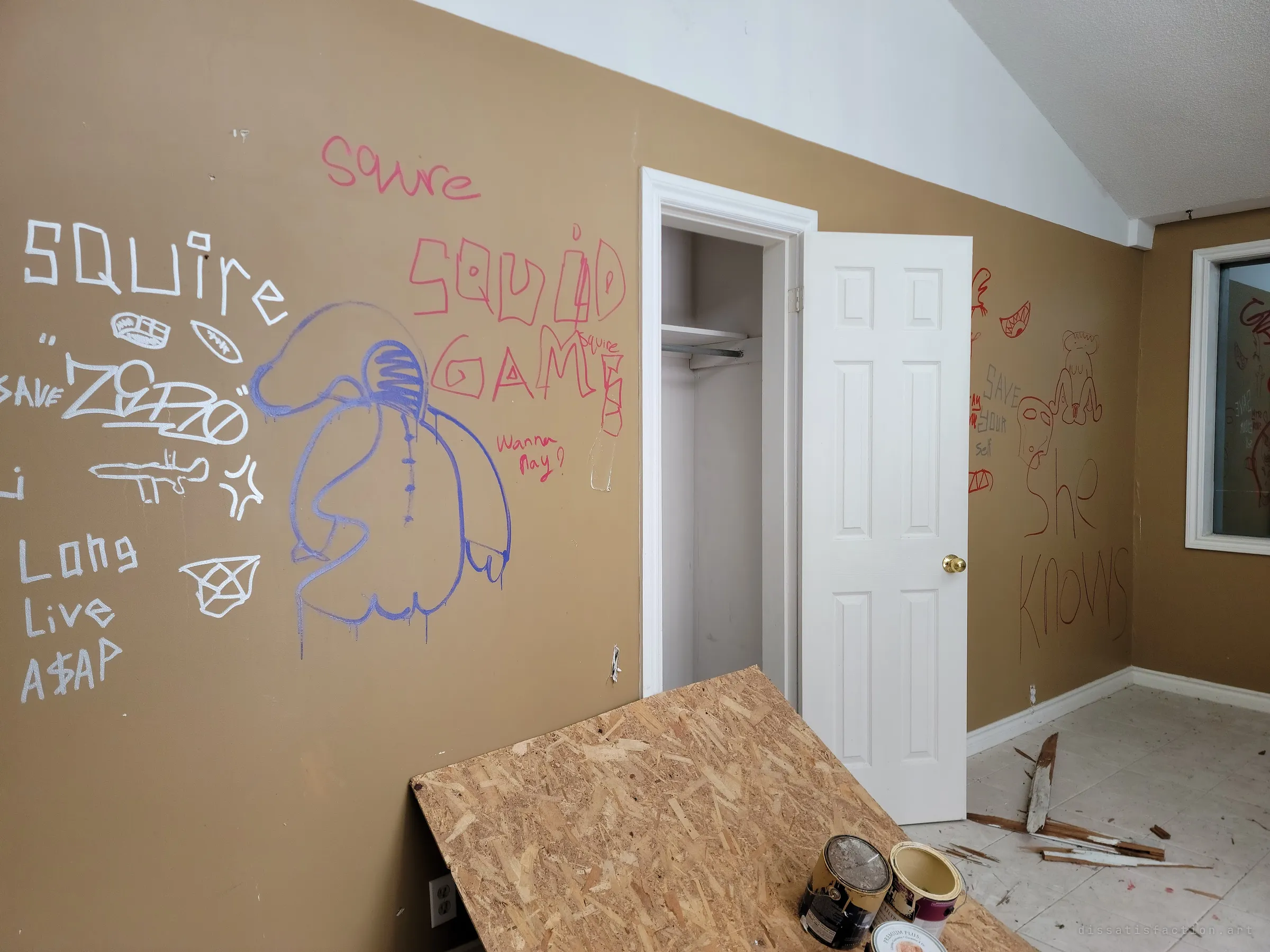 A bedroom with a vaulted ceiling, its tan walls covered in marker and spray-paint graffiti around an open closet door, with a sheet of OSB and paint cans in the foreground. Later visitors have turned the empty room into a sketchbook.