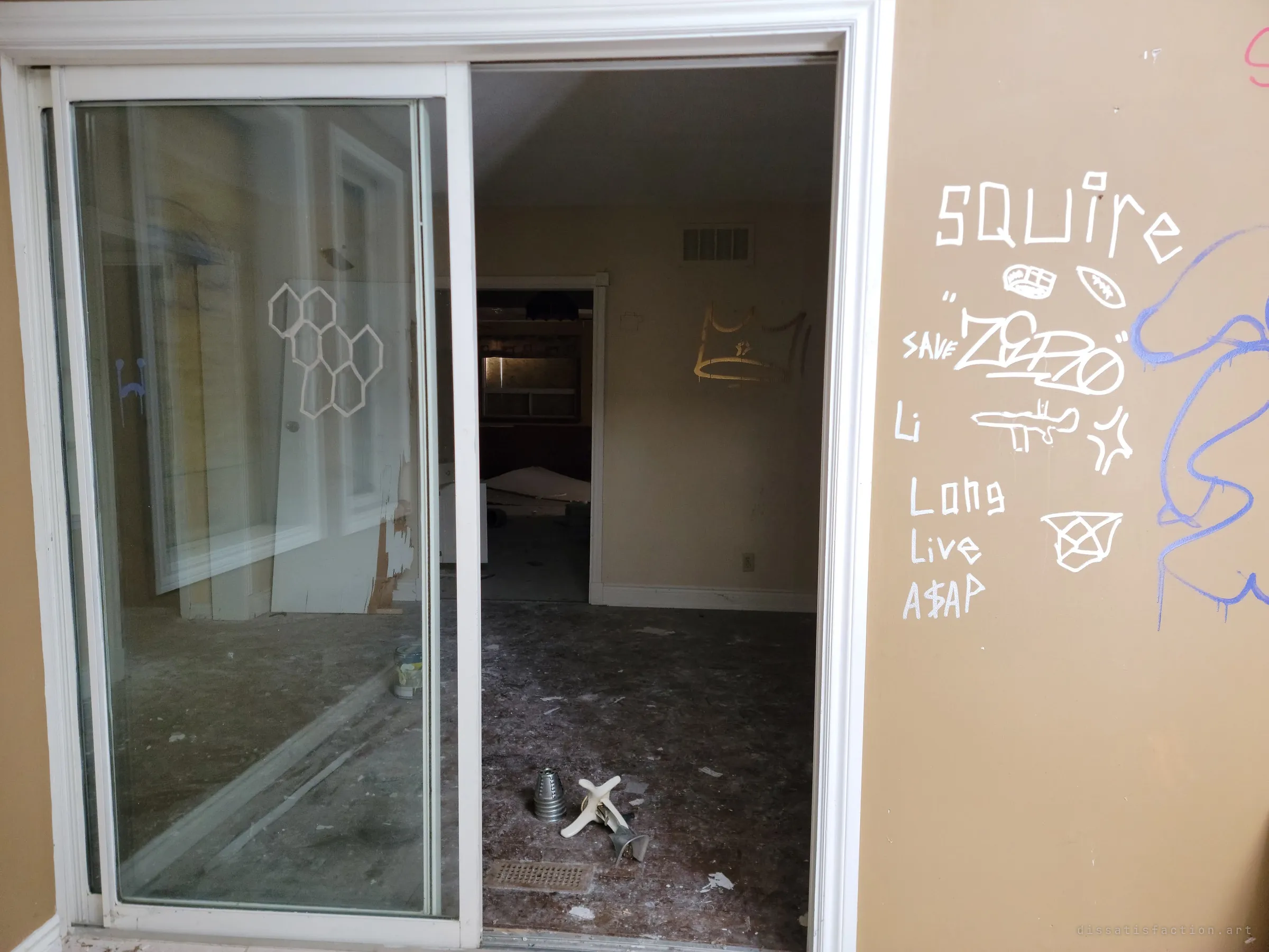 A sliding glass door stands half open onto a dim, debris-strewn room, with a white graffiti tag wall beside it and a gold crown sprayed on the far wall. The glass reflects the boarded sunroom behind the photographer.