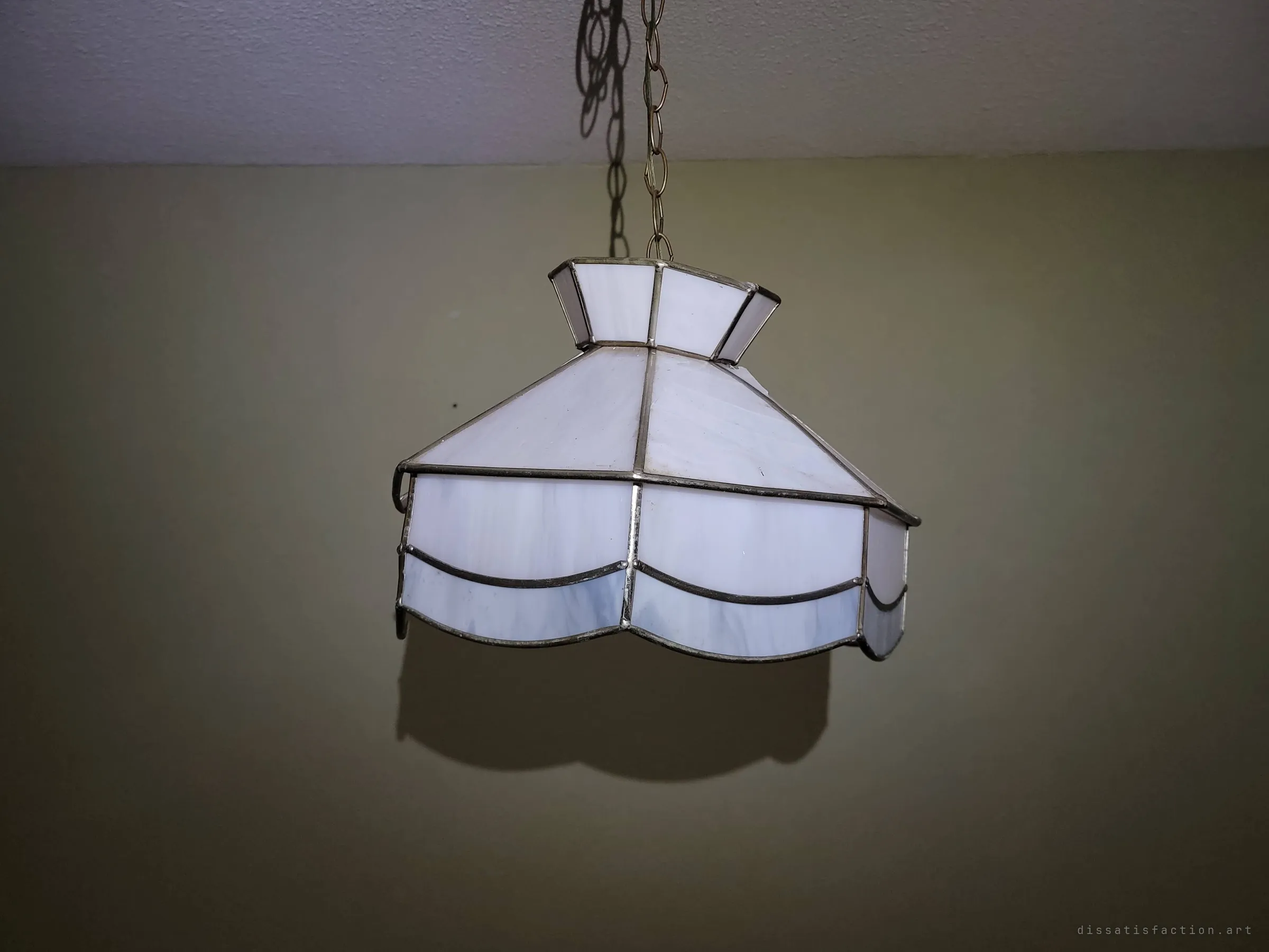 A milky white stained-glass pendant lamp hangs on a chain from a textured ceiling, casting a sharp shadow on the olive wall. A dated domestic detail left untouched in the house.