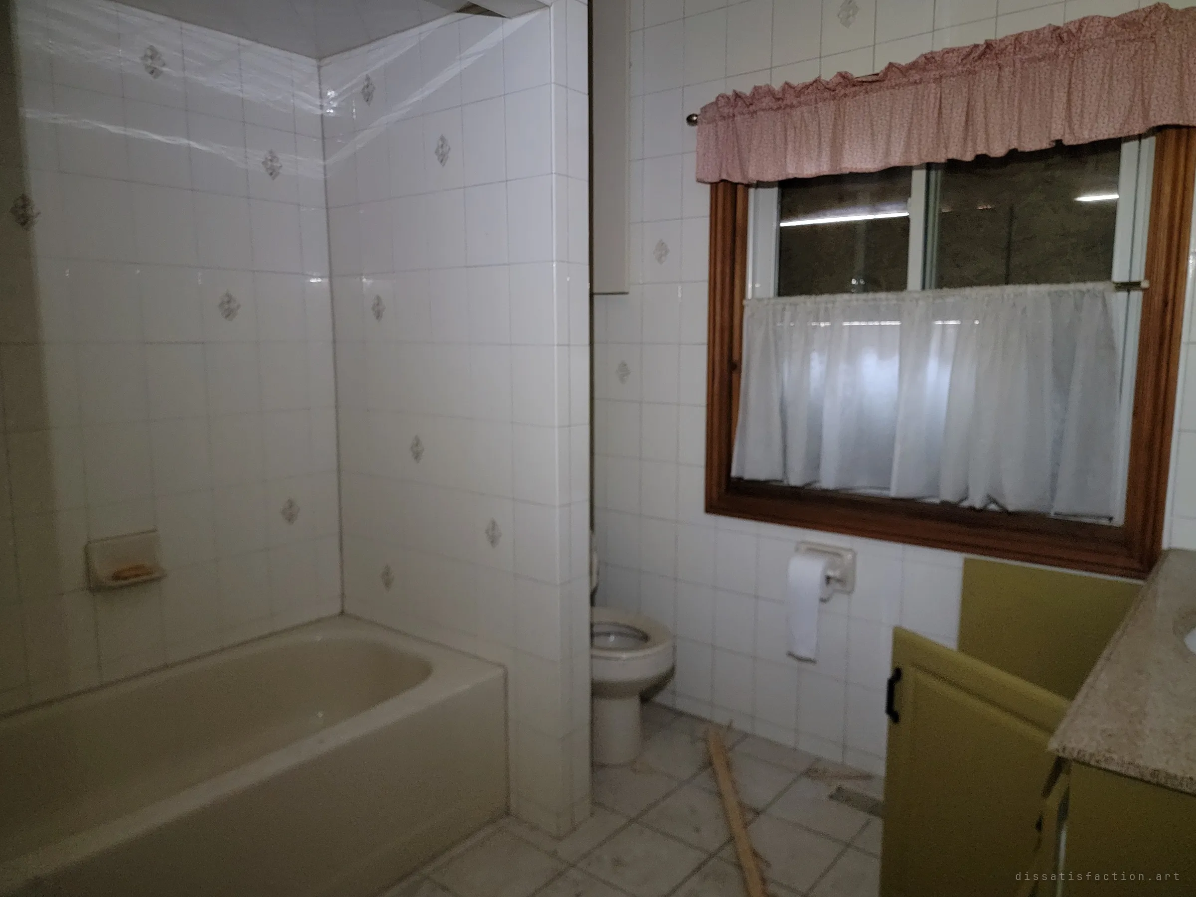 A dim tiled bathroom with a beige tub, toilet and olive vanity under a window dressed in a pink valance and white café curtain. Cobwebs stretch across the shower corner.