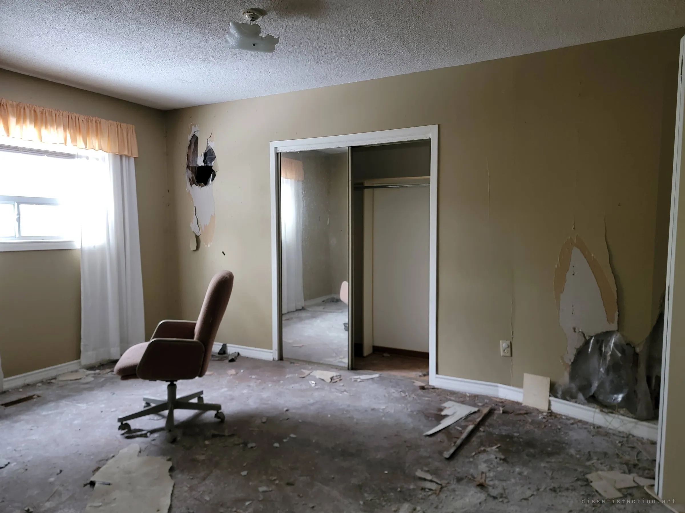 A bedroom with torn drywall and a bare subfloor, where a lone pink office chair faces a mirrored closet door. Soft daylight falls through a curtained window.