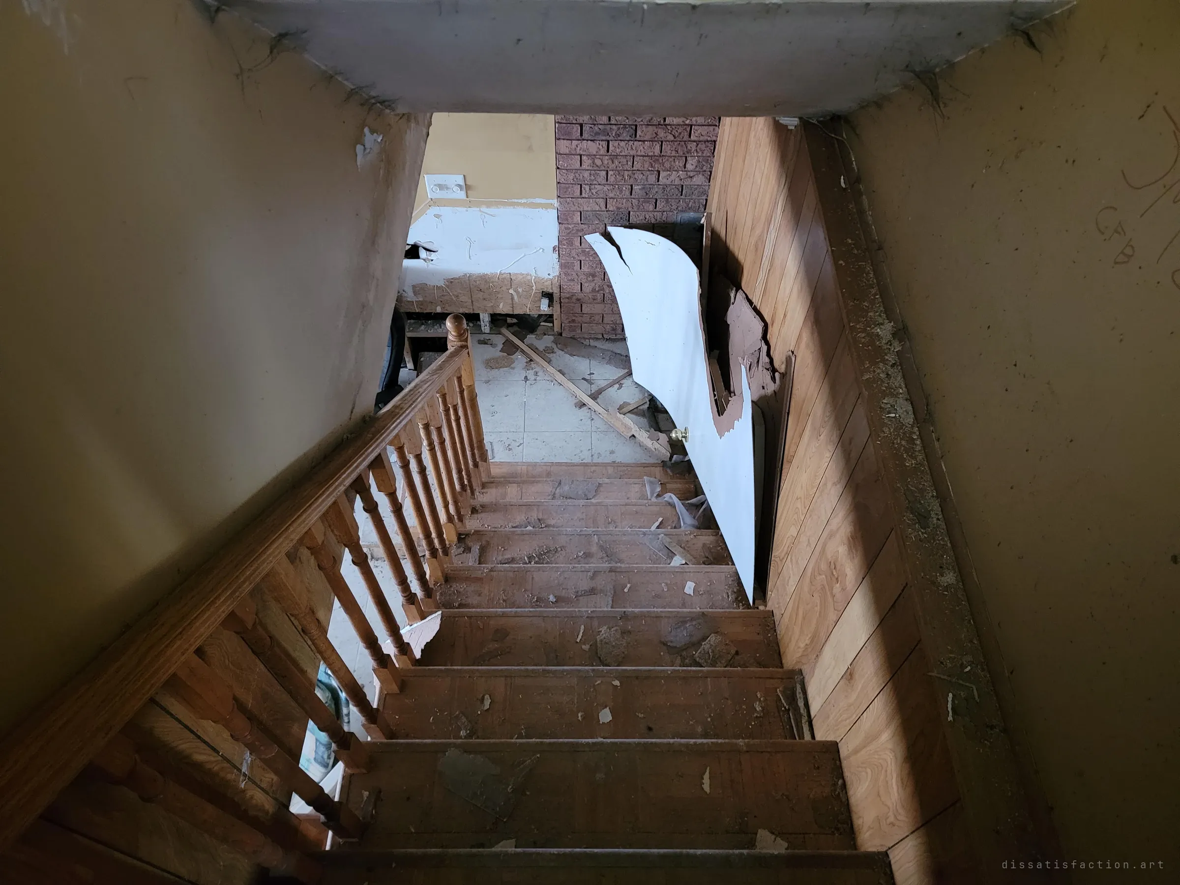 Looking down a wooden staircase with turned balusters, its steps scattered with debris, toward a lower floor with a brick wall and a torn-off door leaning against wood panelling.