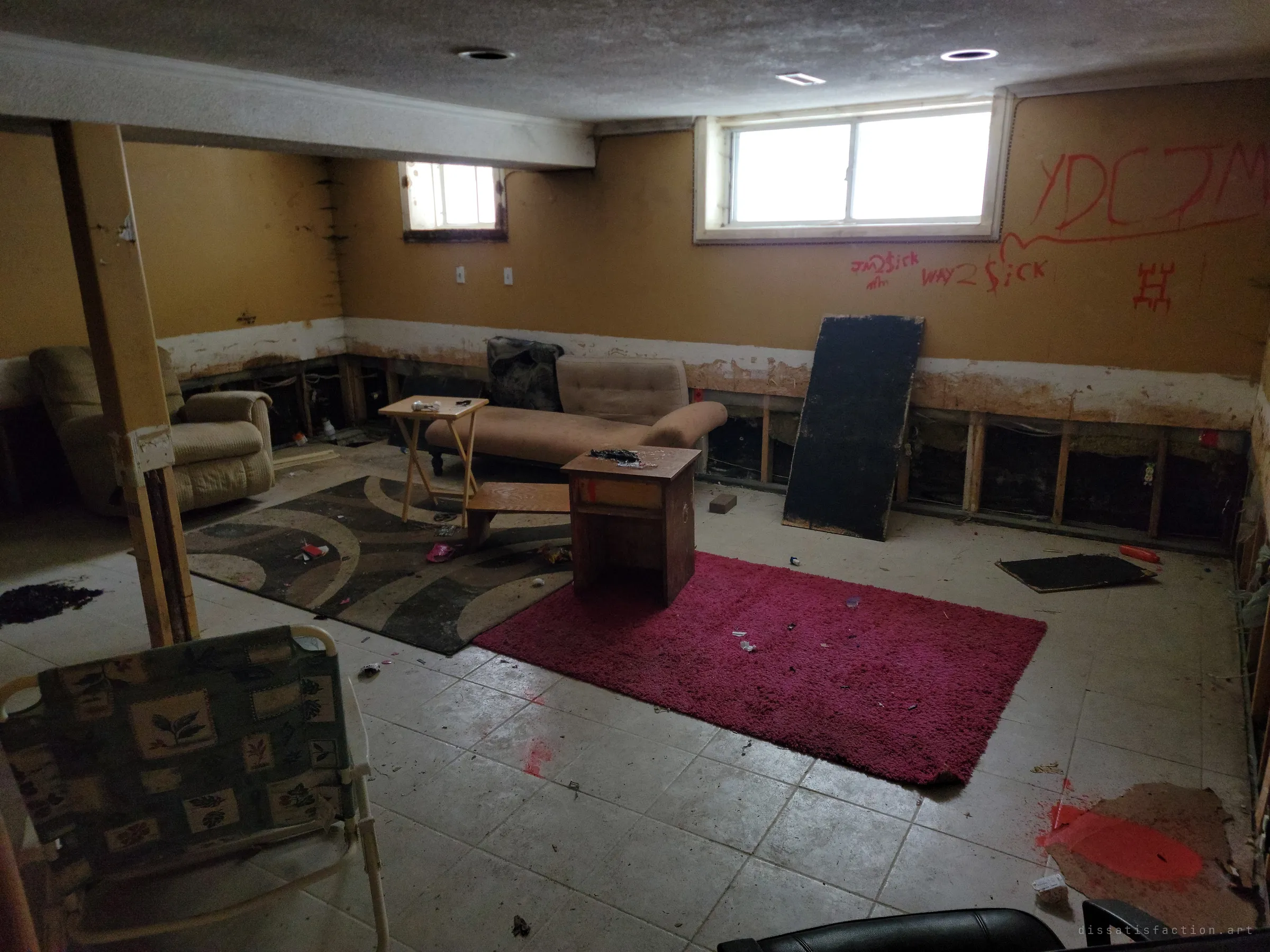 A basement den with its lower drywall cut away after a flood, left with a recliner, a chaise, rugs and scattered junk, and red graffiti across the wall. Small high windows let in flat daylight.