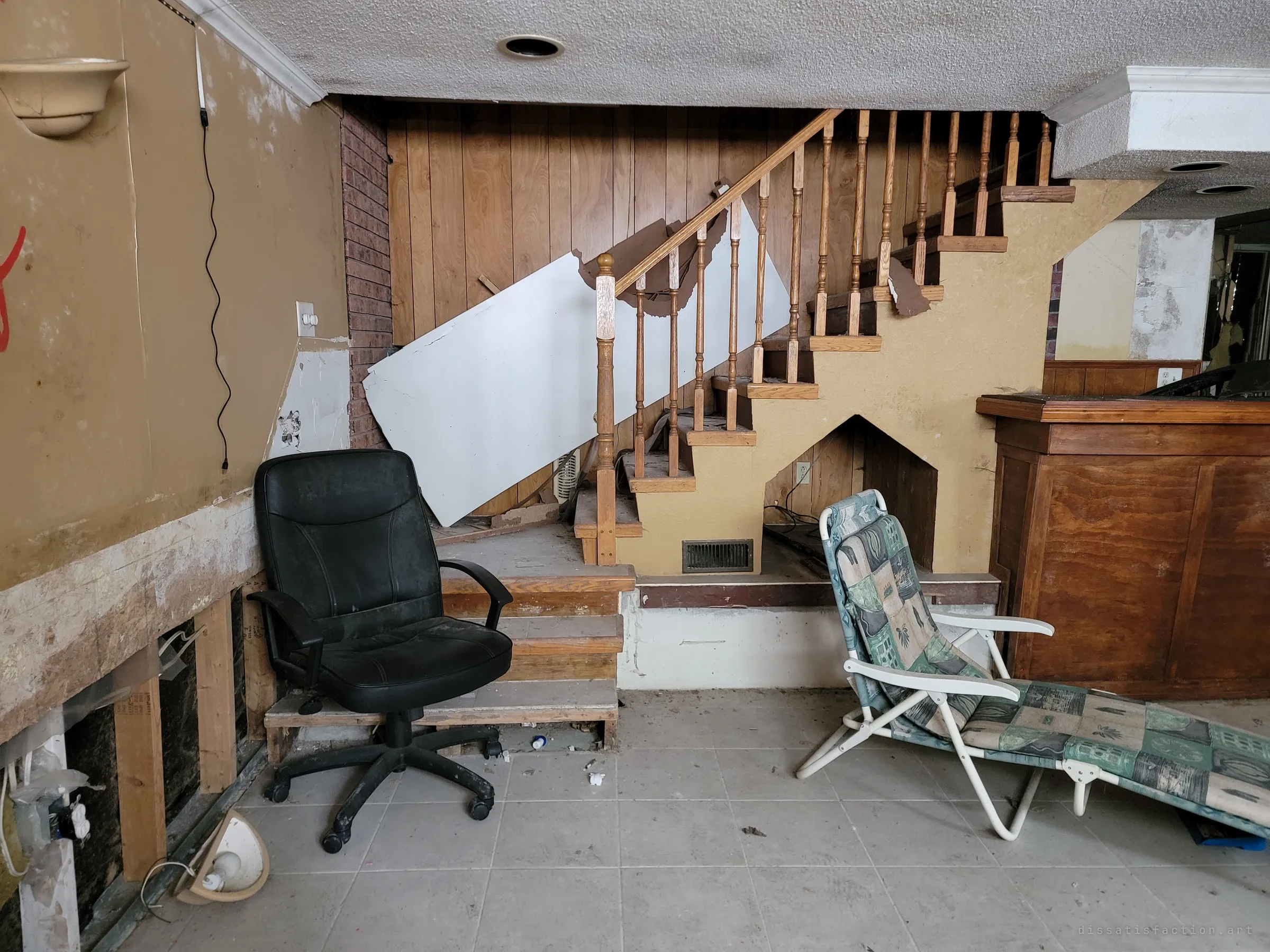 An office chair and a folding lounger sit abandoned at the foot of a wooden basement staircase, a loose sheet of drywall leaning against the banister. Everyday furniture left in a gutted room gives the space an eerie, paused feel.
