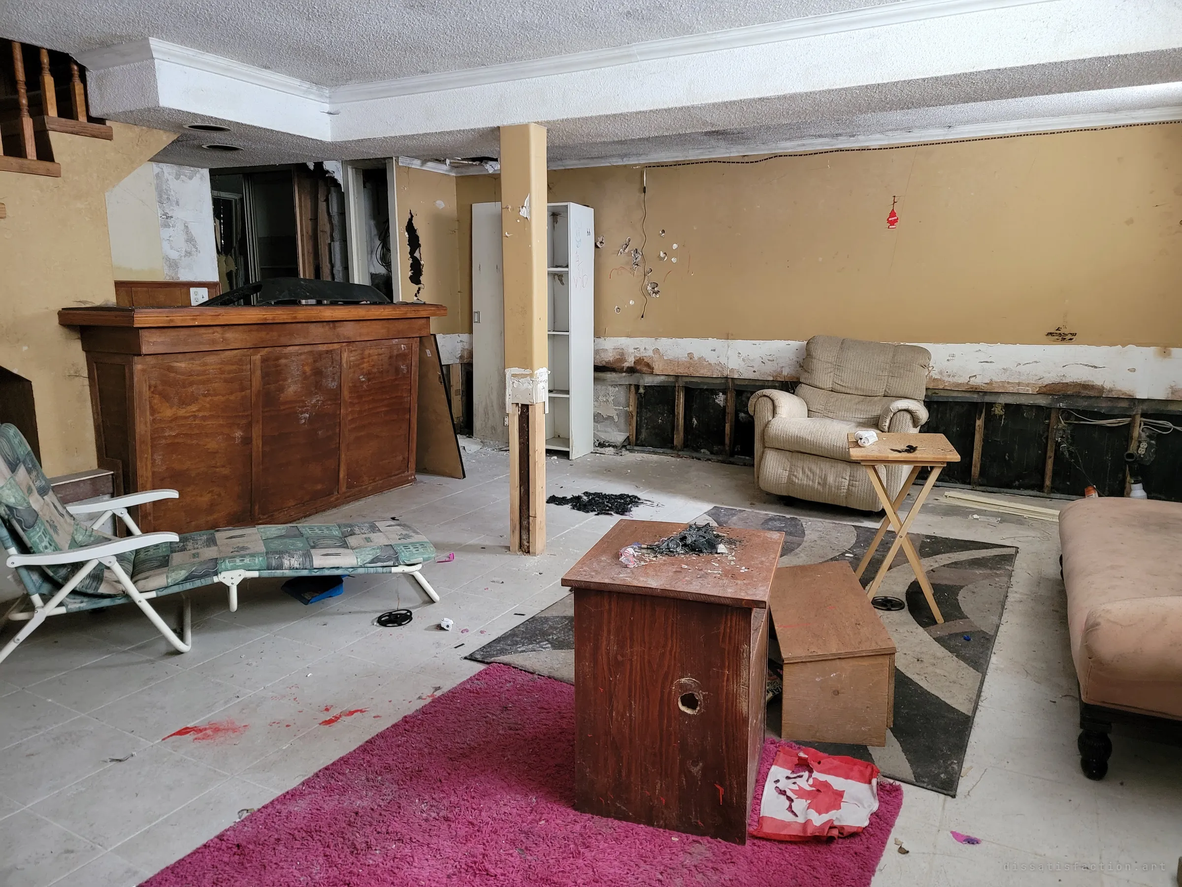 A wide view of the finished basement: a wooden bar, a recliner, a lounger and a scorched crate on a pink rug, with a crumpled national flag on the floor. Burn marks and scattered debris show the space has been used since it was left.