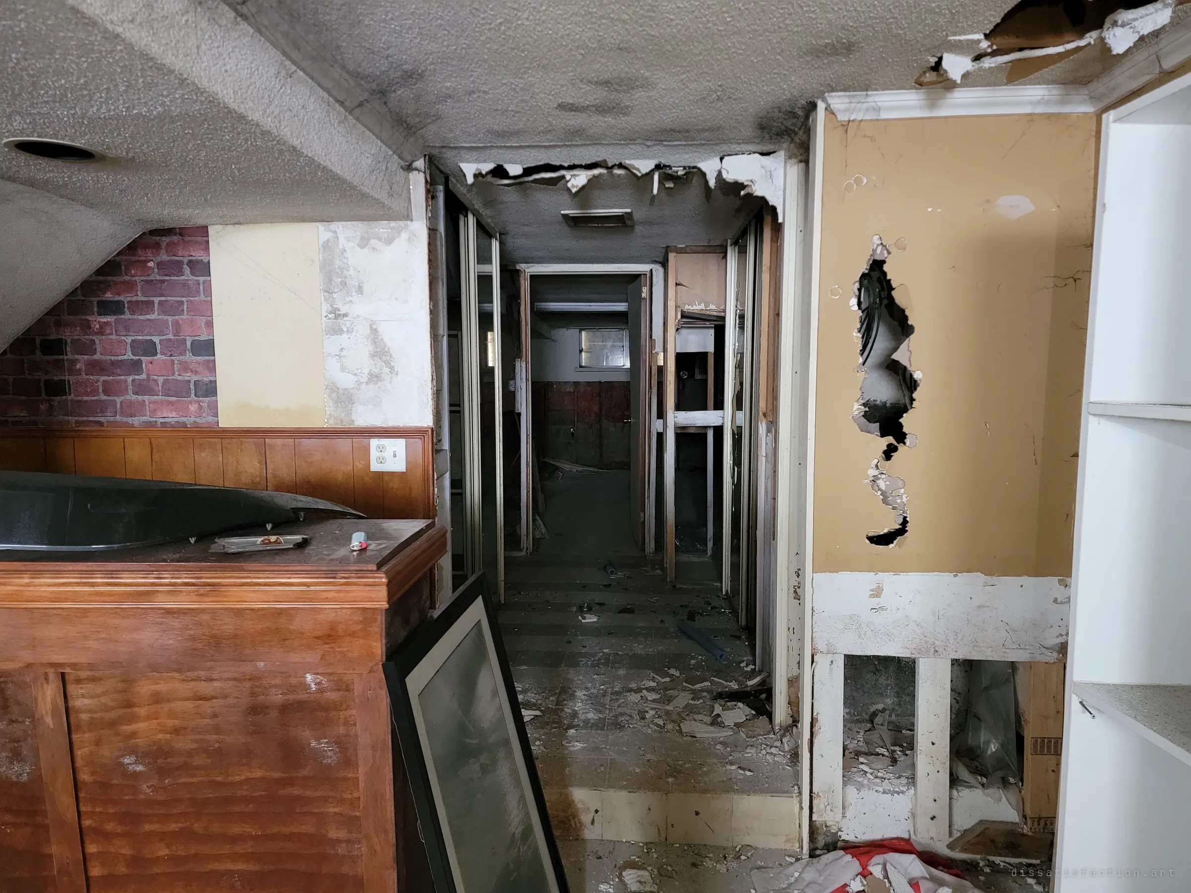 A narrow corridor leads off the basement bar past torn ceiling drywall and a punched-through wall toward darker back rooms. The framed picture leaning on the bar hints at what was left behind.