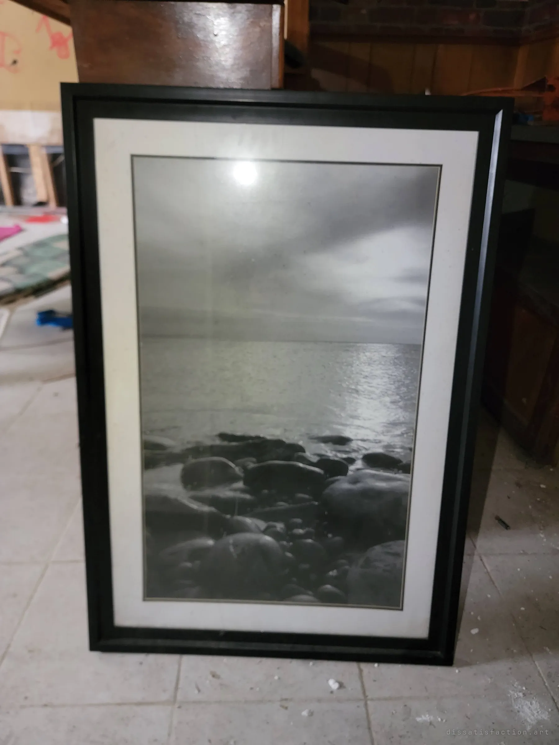 A framed black-and-white print of a calm, rocky shoreline leans against the bar on the dusty tile floor. A quiet decorative piece that outlasted the rest of the room.