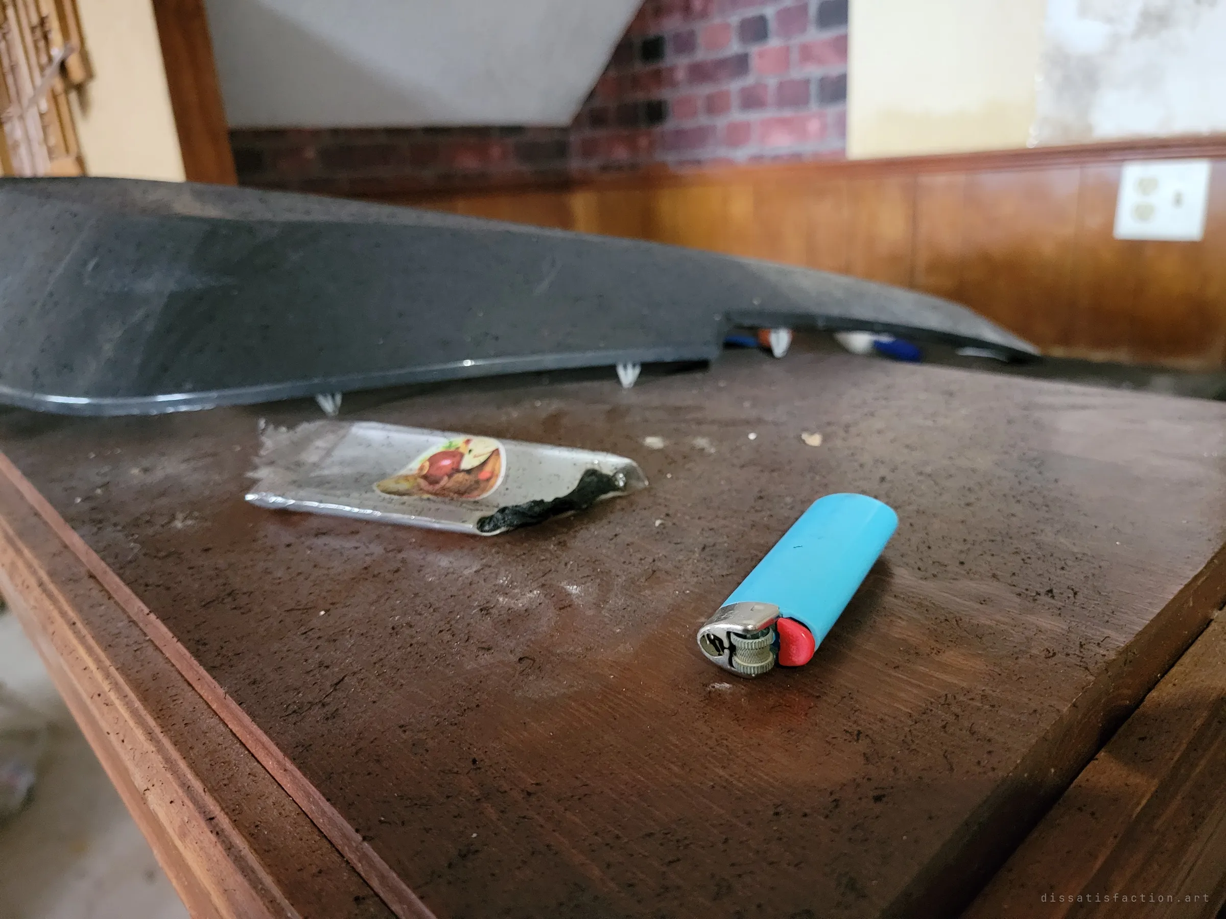 Close-up of a blue lighter and a scorched strip of foil on the dusty bar top. Small traces of the people who passed through after the house was abandoned.