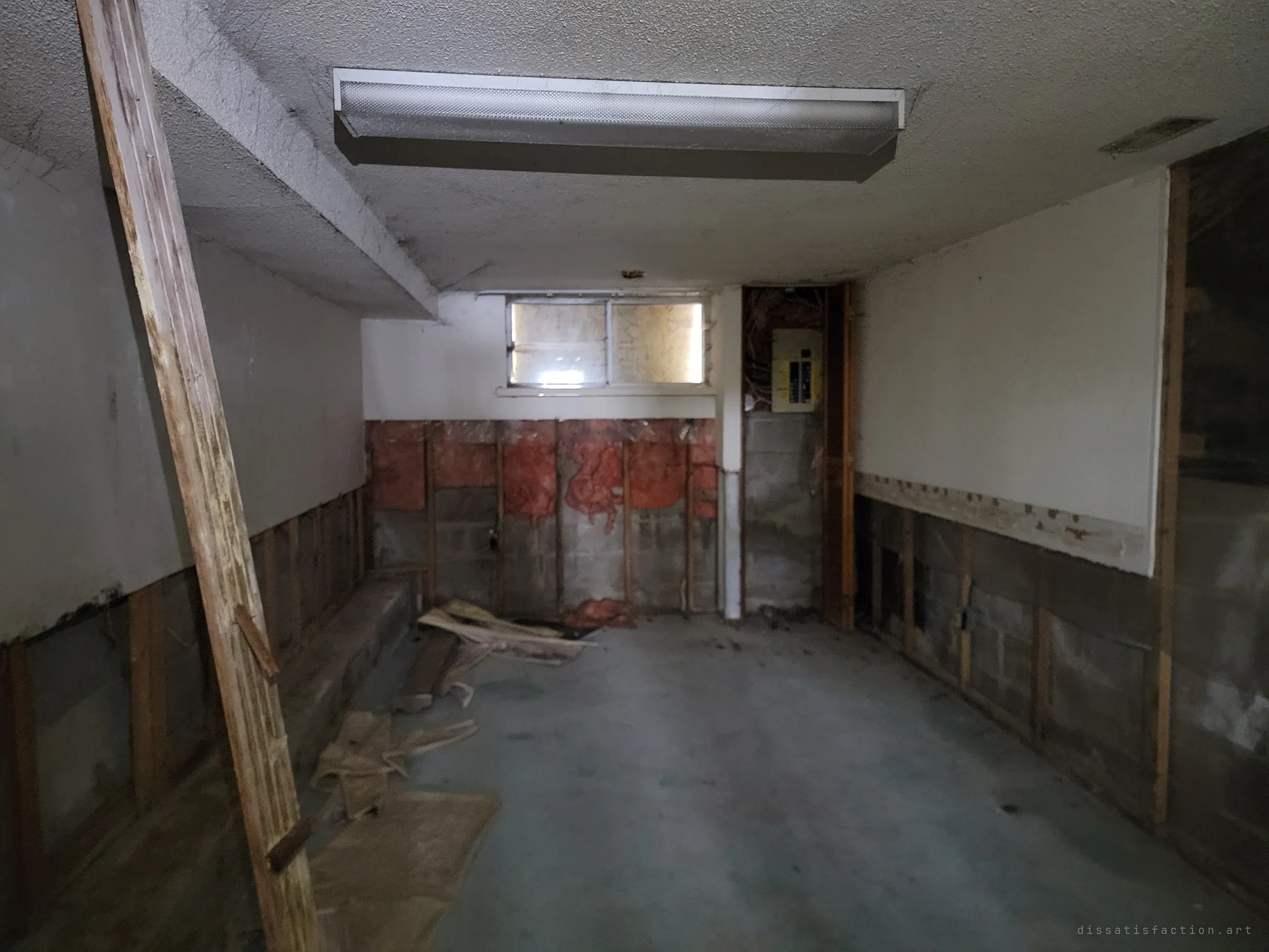 A dim back room of the basement with a fluorescent tube light, a boarded window and the walls cut open to insulation and cinder block. The open electrical panel and bare studs show the renovation that stopped midway.