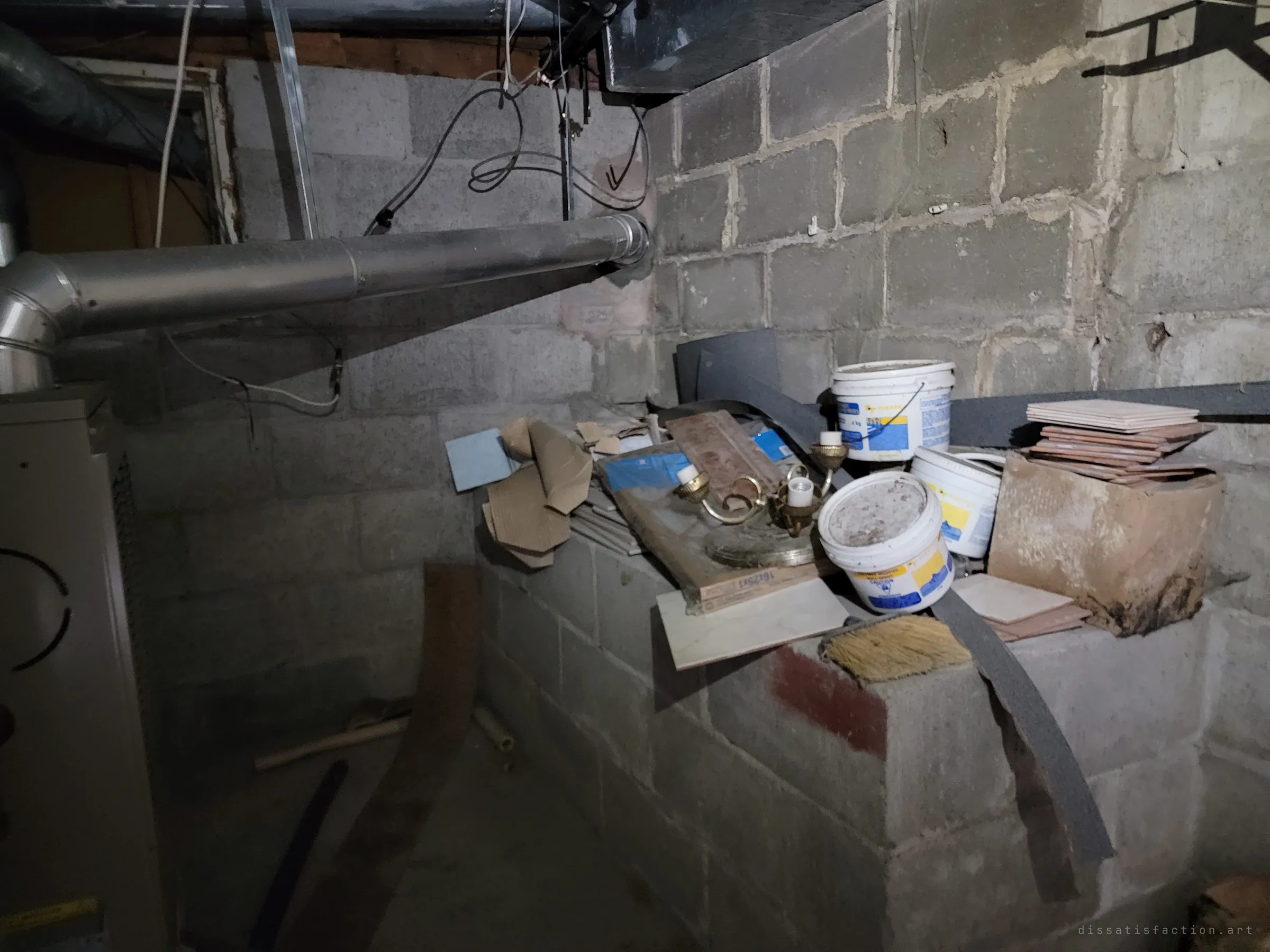 A cinder-block utility corner by the furnace, its ledge piled with tile offcuts, old paint pails and a detached light fixture. Leftover supplies from repairs that were never finished.