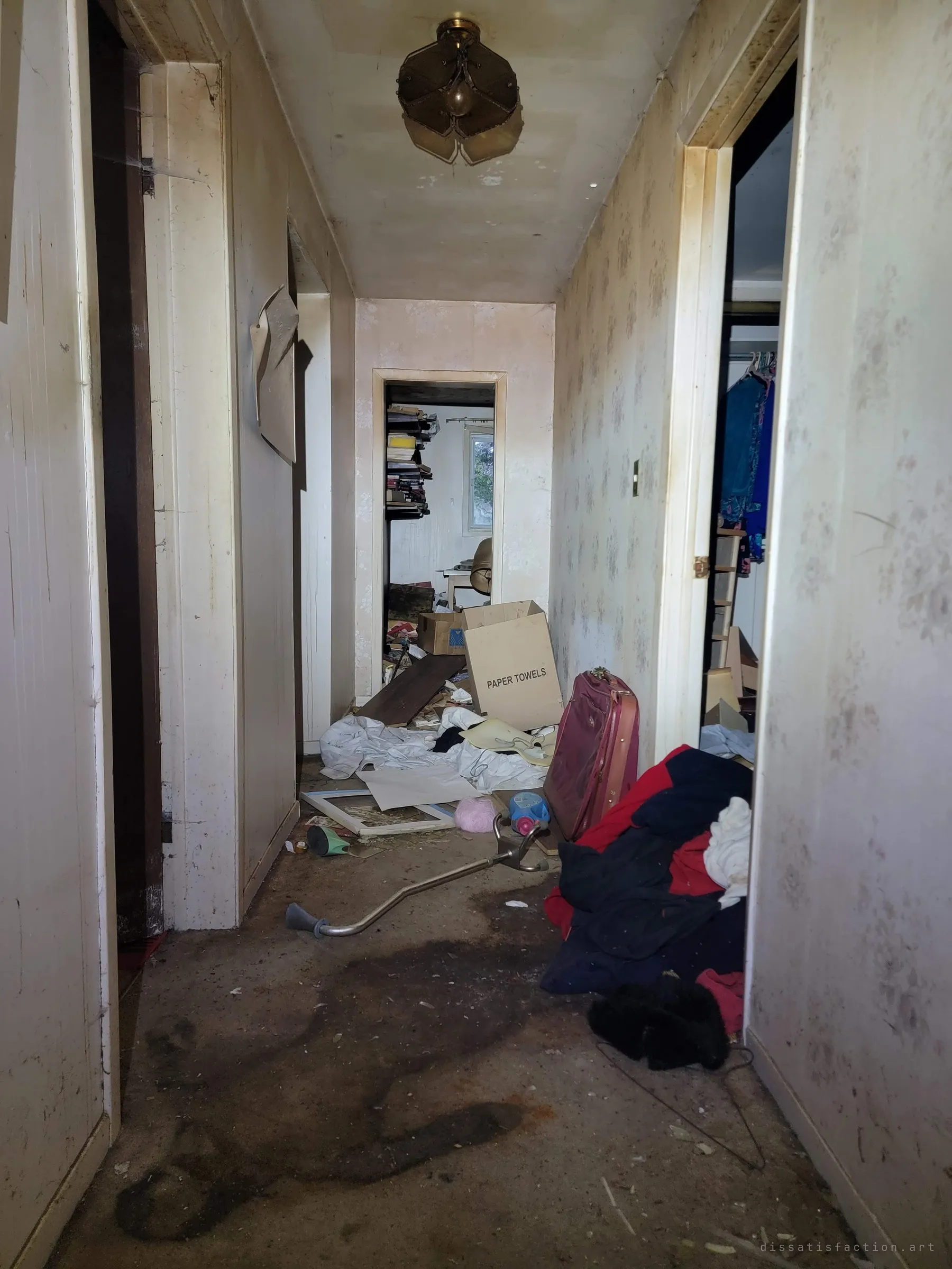 A stained hallway runs toward a book-lined room, its floor littered with clothes, a suitcase and a dropped forearm crutch.