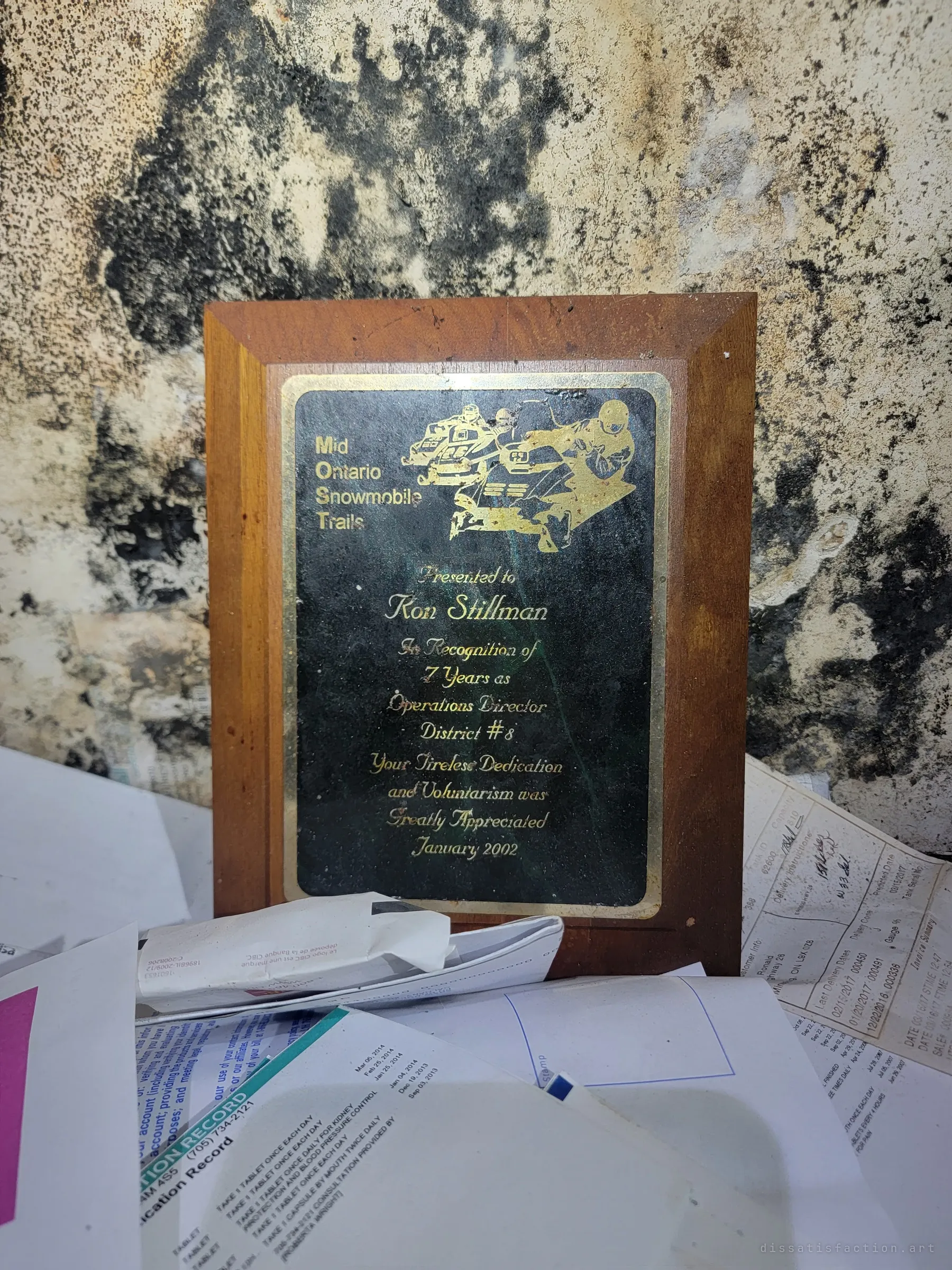 A volunteer service plaque rests against a mould-blackened wall, surrounded by medication records and old bills left behind.