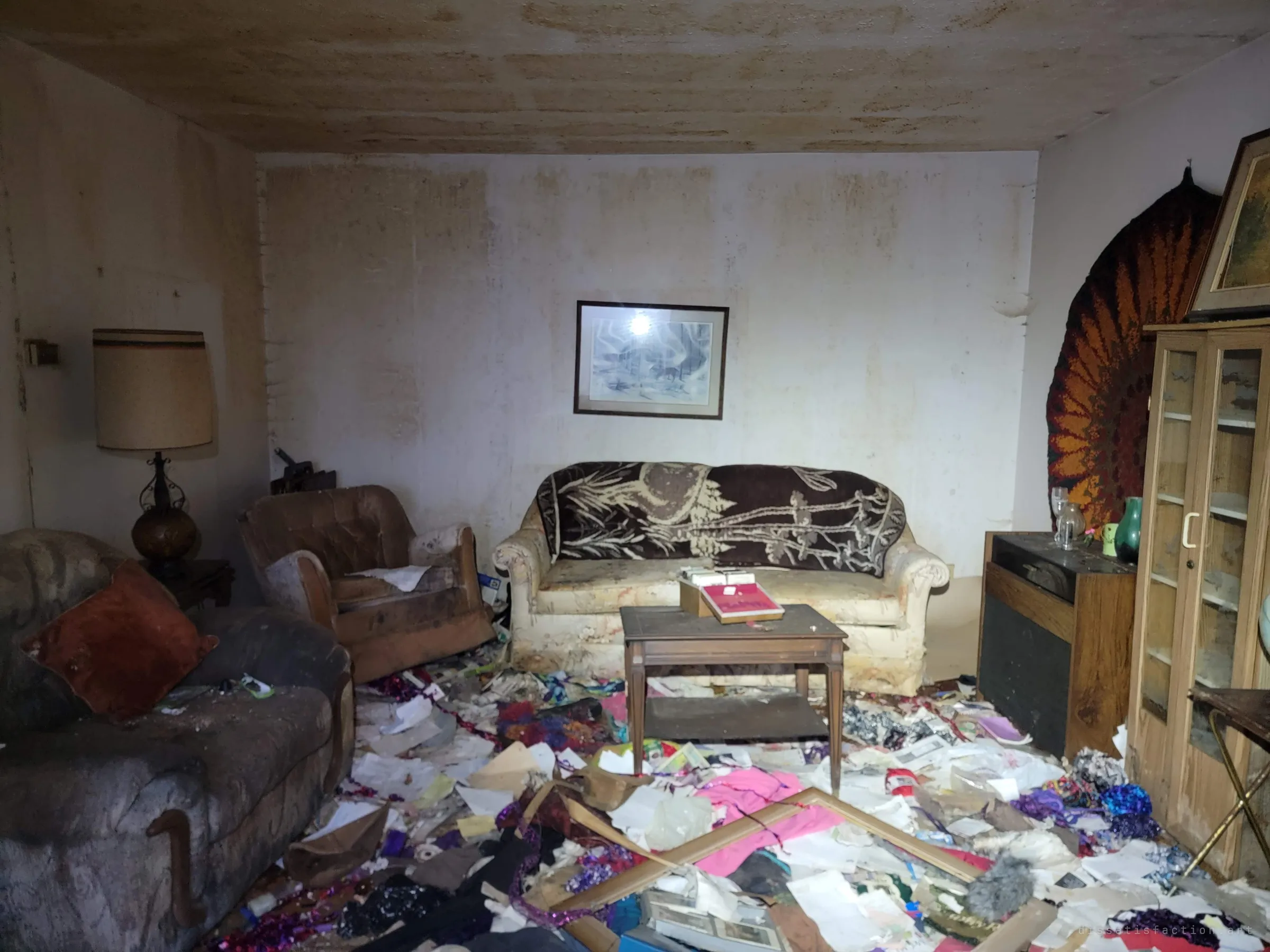 A water-stained living room with a floral-draped sofa and a framed winter print, its floor buried in papers, tinsel and fabric.