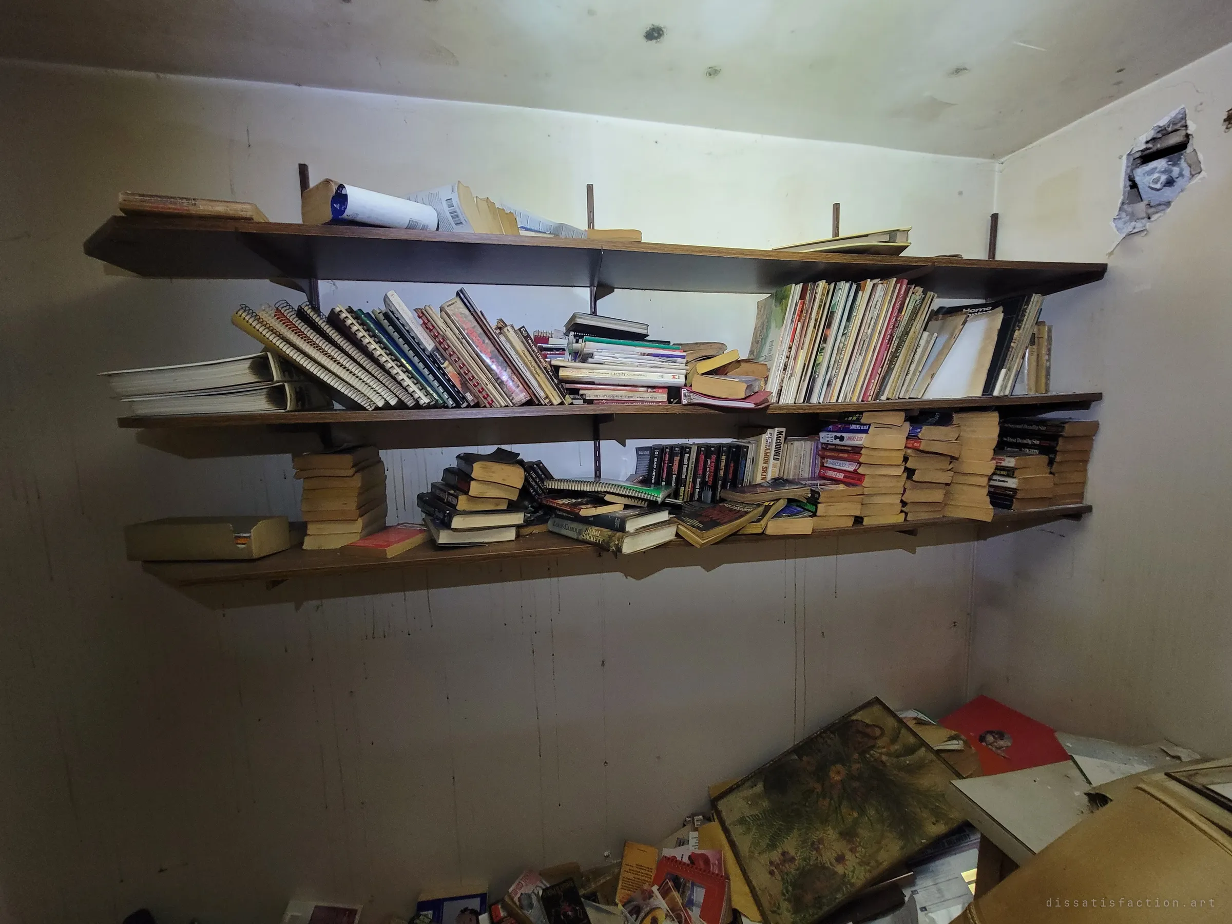 Sagging wall shelves still packed with spiral cookbooks, magazines and stacks of yellowed paperbacks, with framed pictures and papers heaped on the floor below. A reading life left exactly where it stopped.