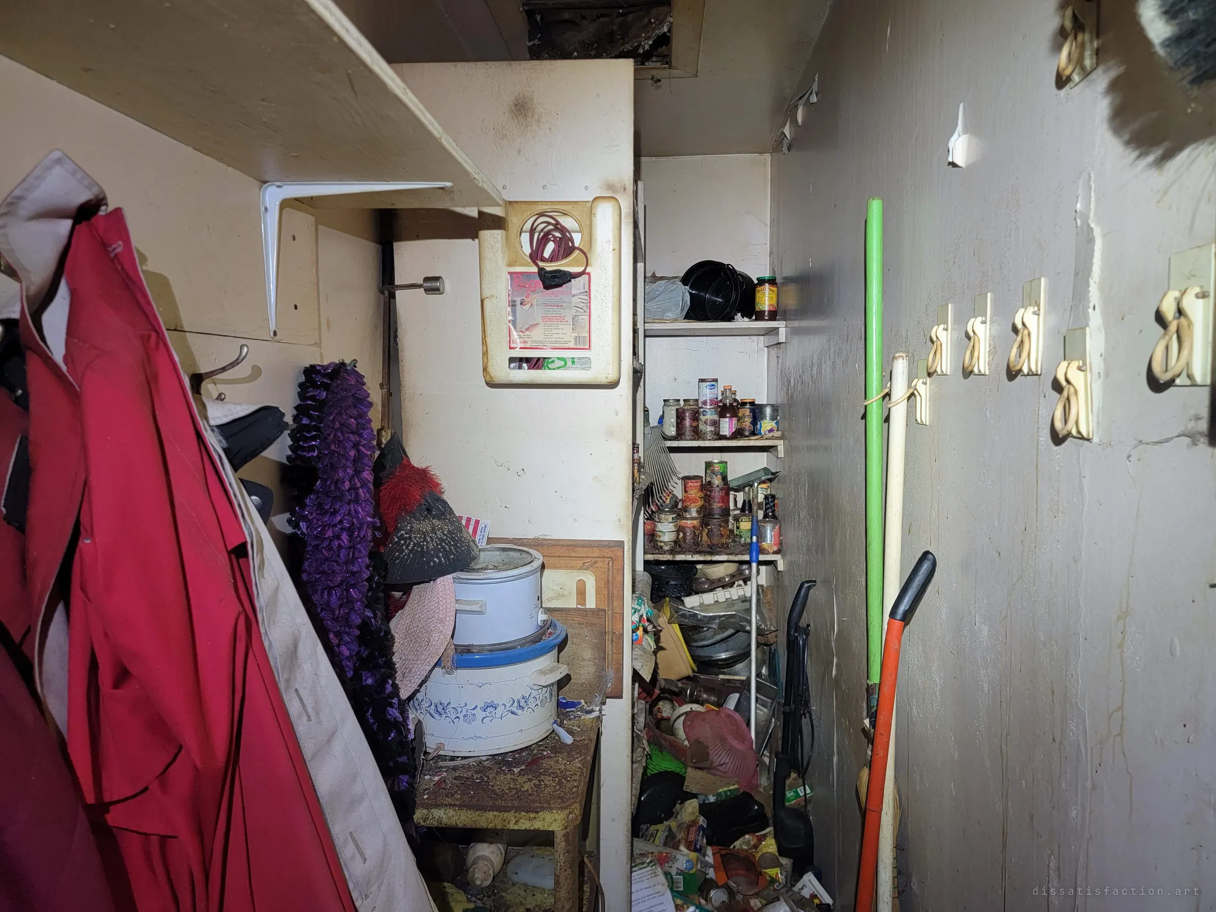 A cramped utility closet: red coats on hooks, slow cookers on a rusted stand, a pantry of canned goods and brooms leaning against a stained wall. Everyday supplies abandoned mid-use.
