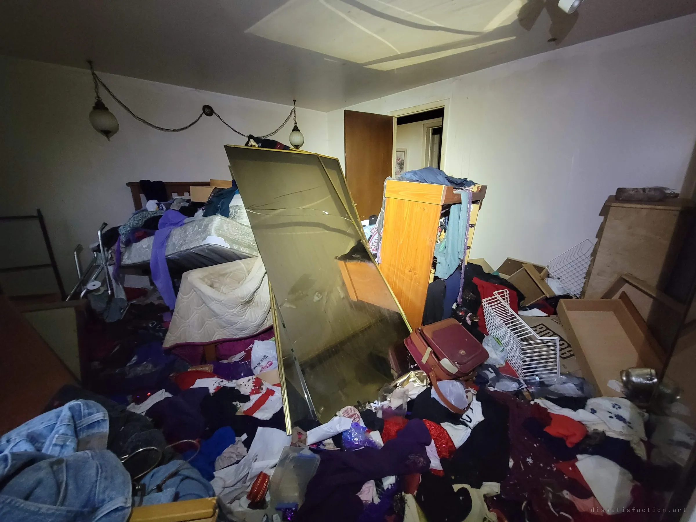A bedroom buried in clothing, with a cracked mirrored door leaning across the mattress and an open dresser behind it, under a flashlight beam. The mess suggests the room was searched or emptied in a hurry.