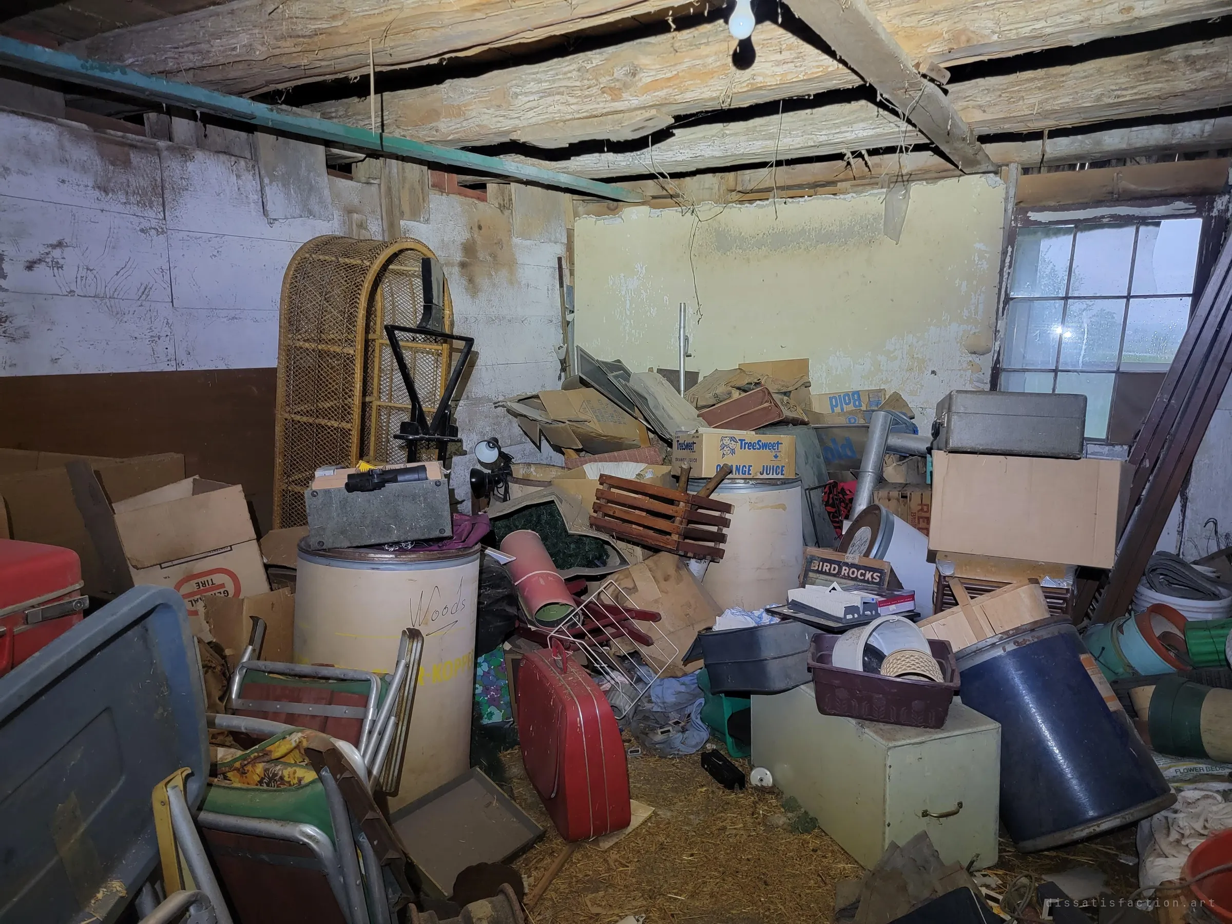 A low outbuilding under rough log joists, crammed with fibre drums, old boxes, a wicker chair, folding lawn chairs and a red suitcase, with a rain-streaked window onto green fields. Decades of storage left to the damp.