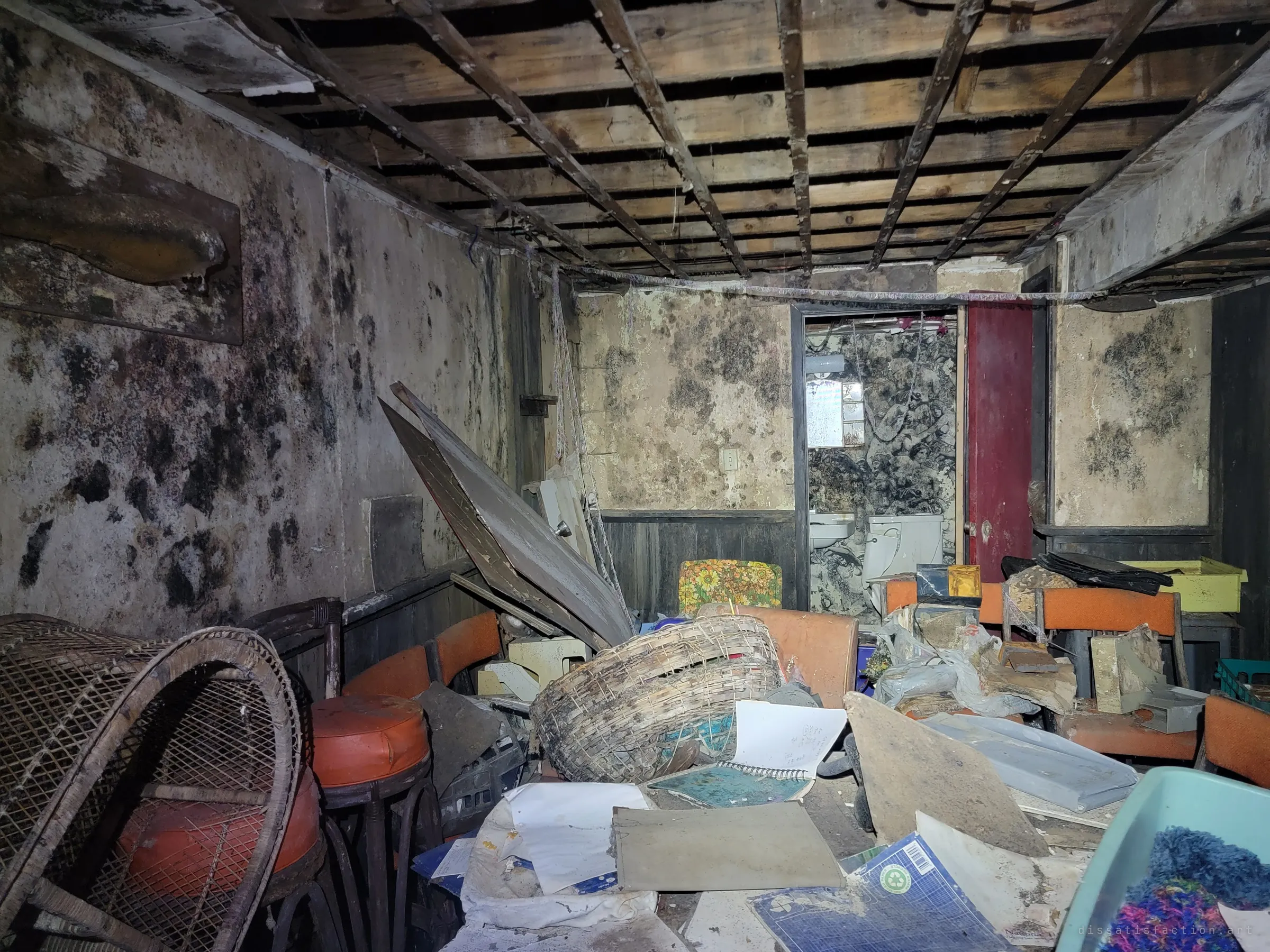A basement rec room with its ceiling stripped to the lath and walls blotched black with mould, orange bar stools and a wicker basket amid scattered notebooks. A red door opens onto an equally mouldy bathroom.