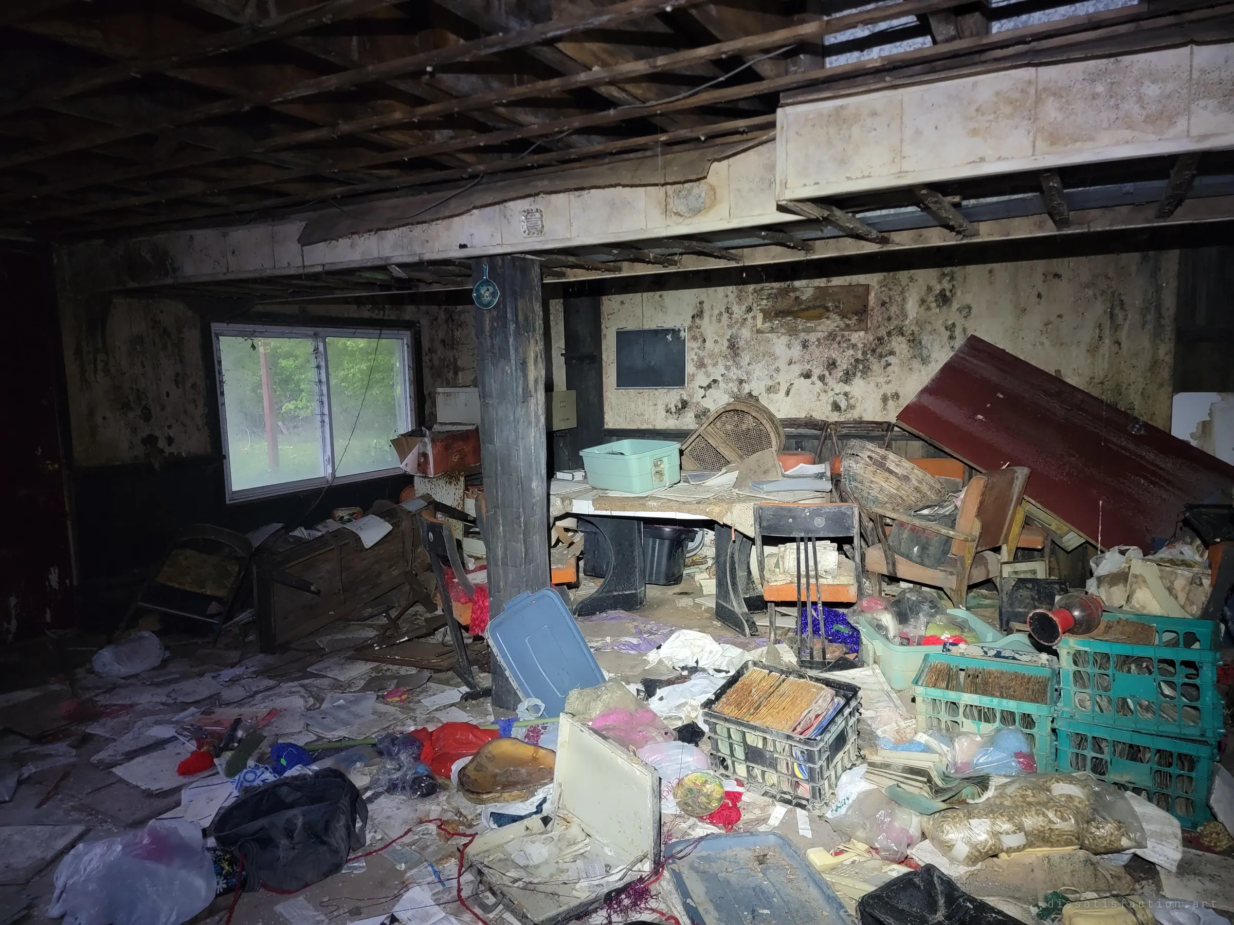 The wider basement: a mould-covered room with the ceiling torn open, a toppled door, crates of old records and papers strewn across the floor, and daylight through a window onto trees. The damage shows how long water has been getting in.
