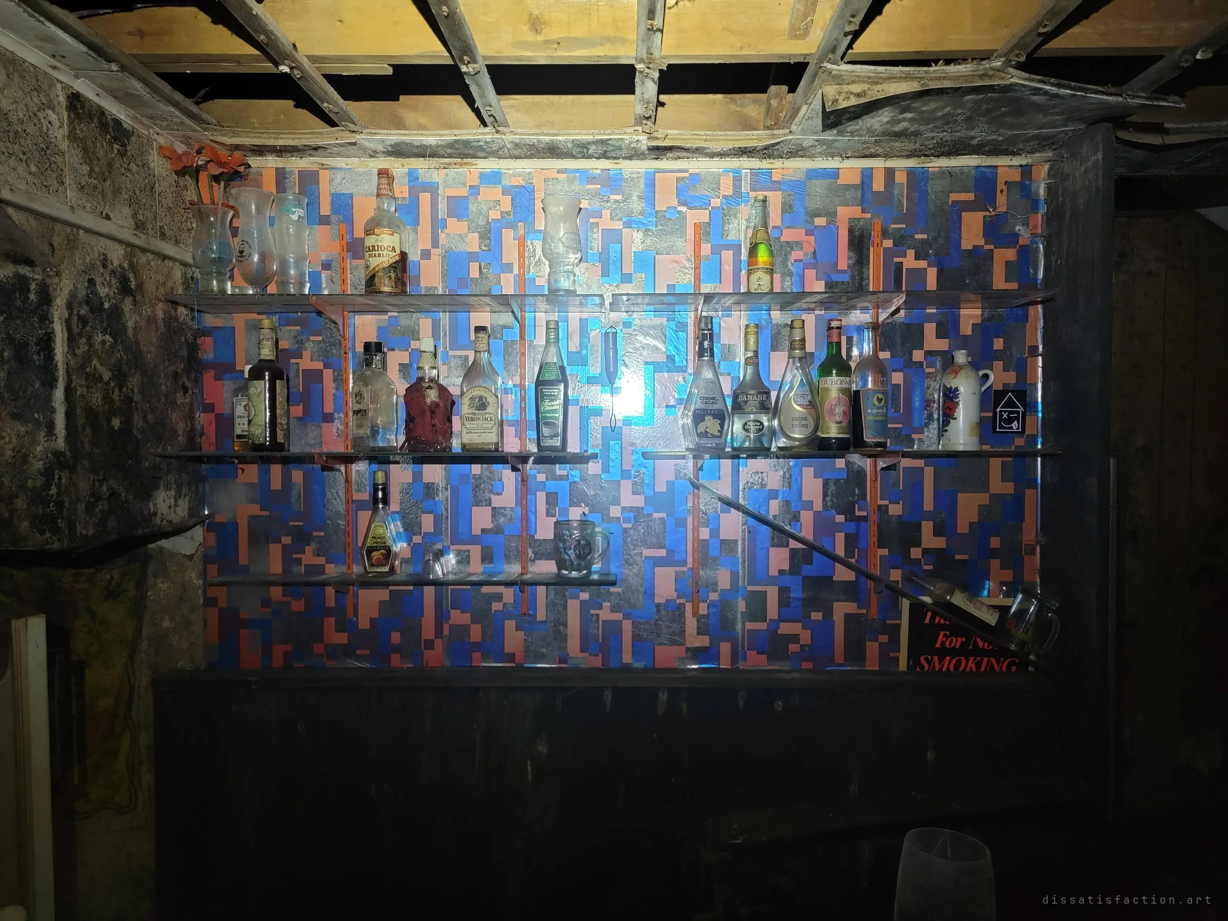A home bar's back wall papered in blue and orange pixel blocks, glass shelves still lined with dusty liqueur bottles and hurricane glasses. A party room frozen in another era.