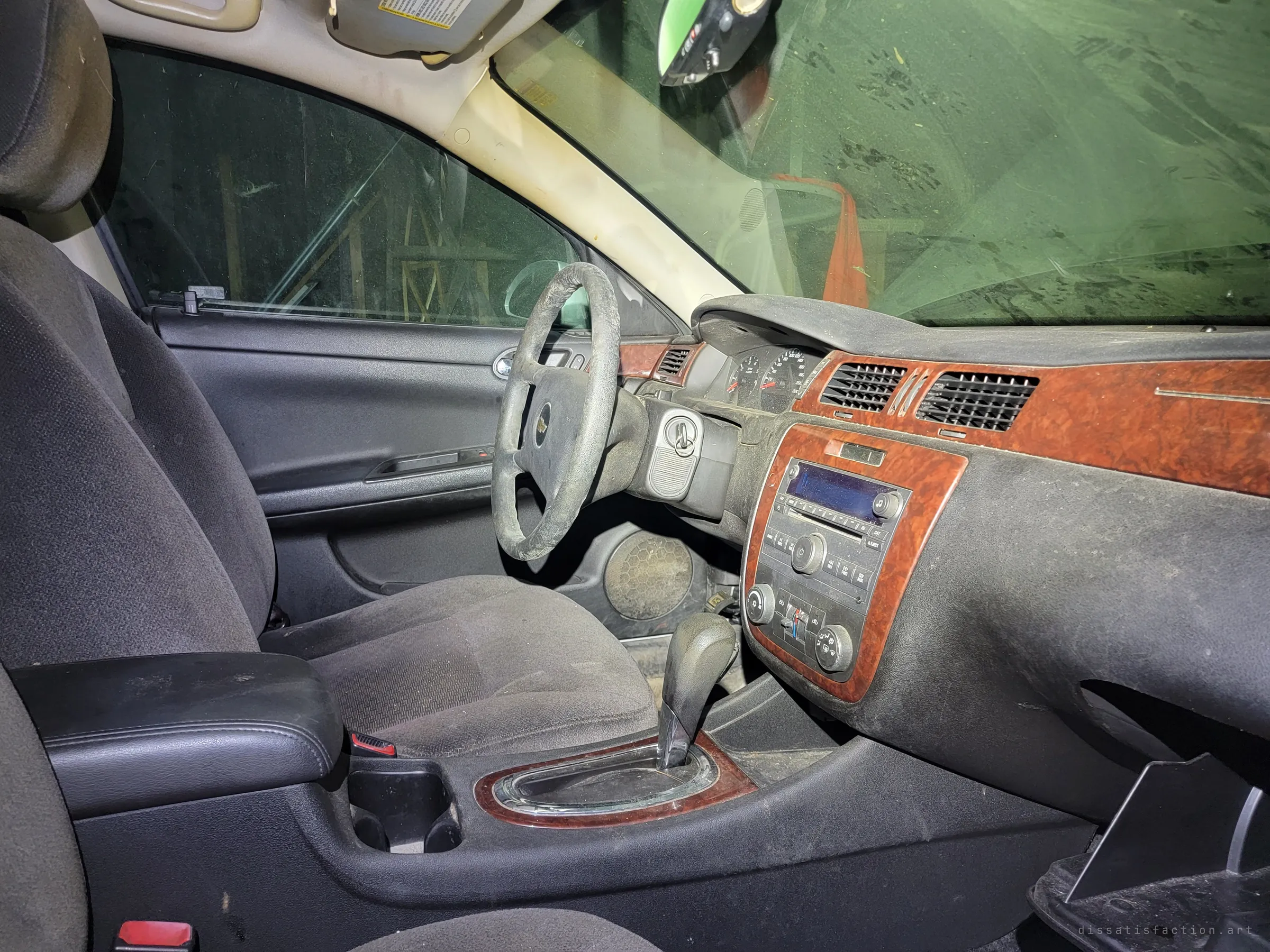 Inside a dust-coated sedan: grimy steering wheel, woodgrain dash and cloth seats, with the garage clutter visible through the windows. A car parked and never driven again.