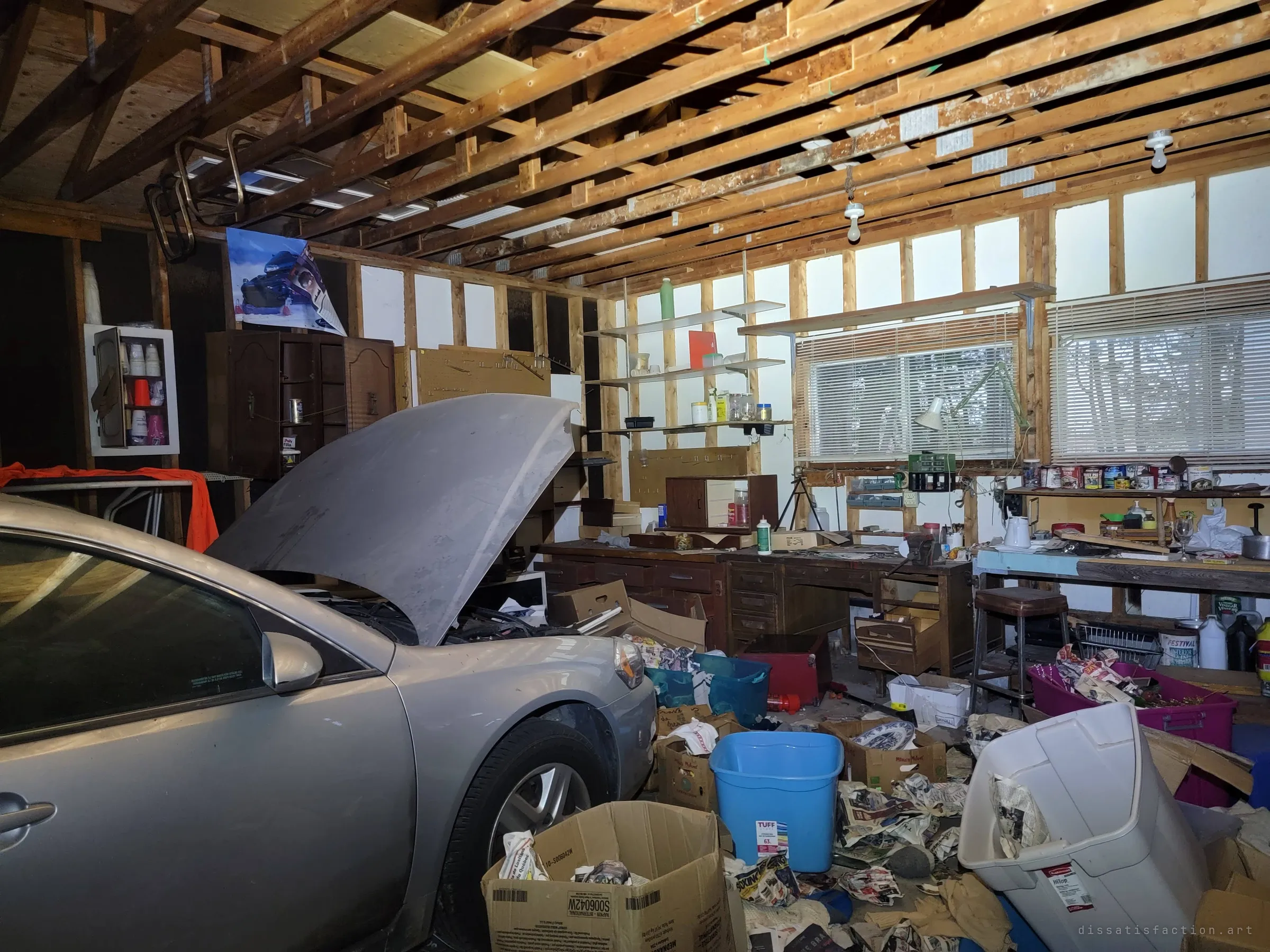 A garage workshop with bare framed walls, the silver sedan's hood propped open, a workbench of paint tins and a floor of torn newspapers and bins. Work left half finished.