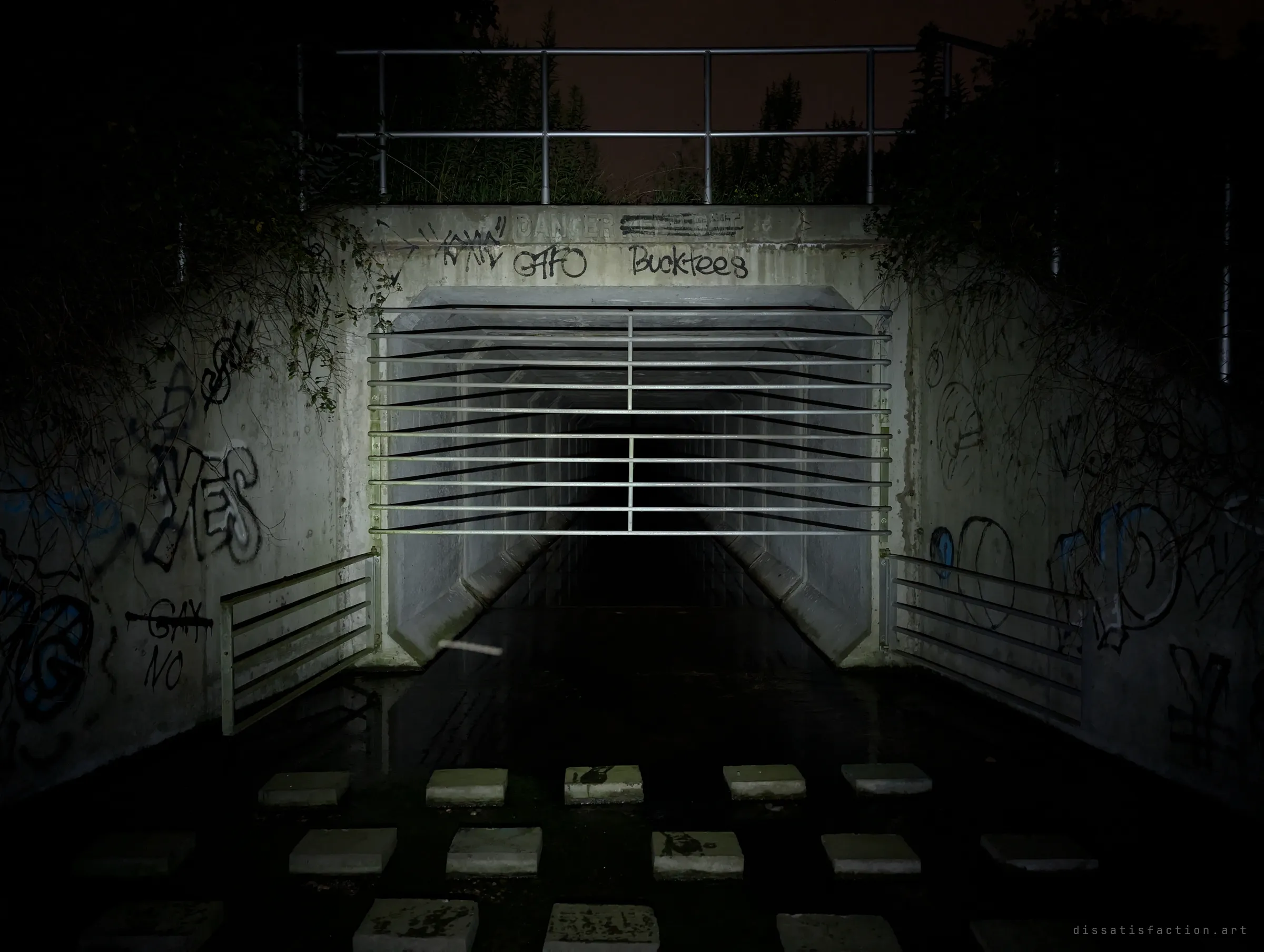 A barred concrete box-culvert outfall at night, its graffiti-covered headwall and faded danger stencil lit by a flashlight over a row of stepping stones. The grate marks the way into the drain.