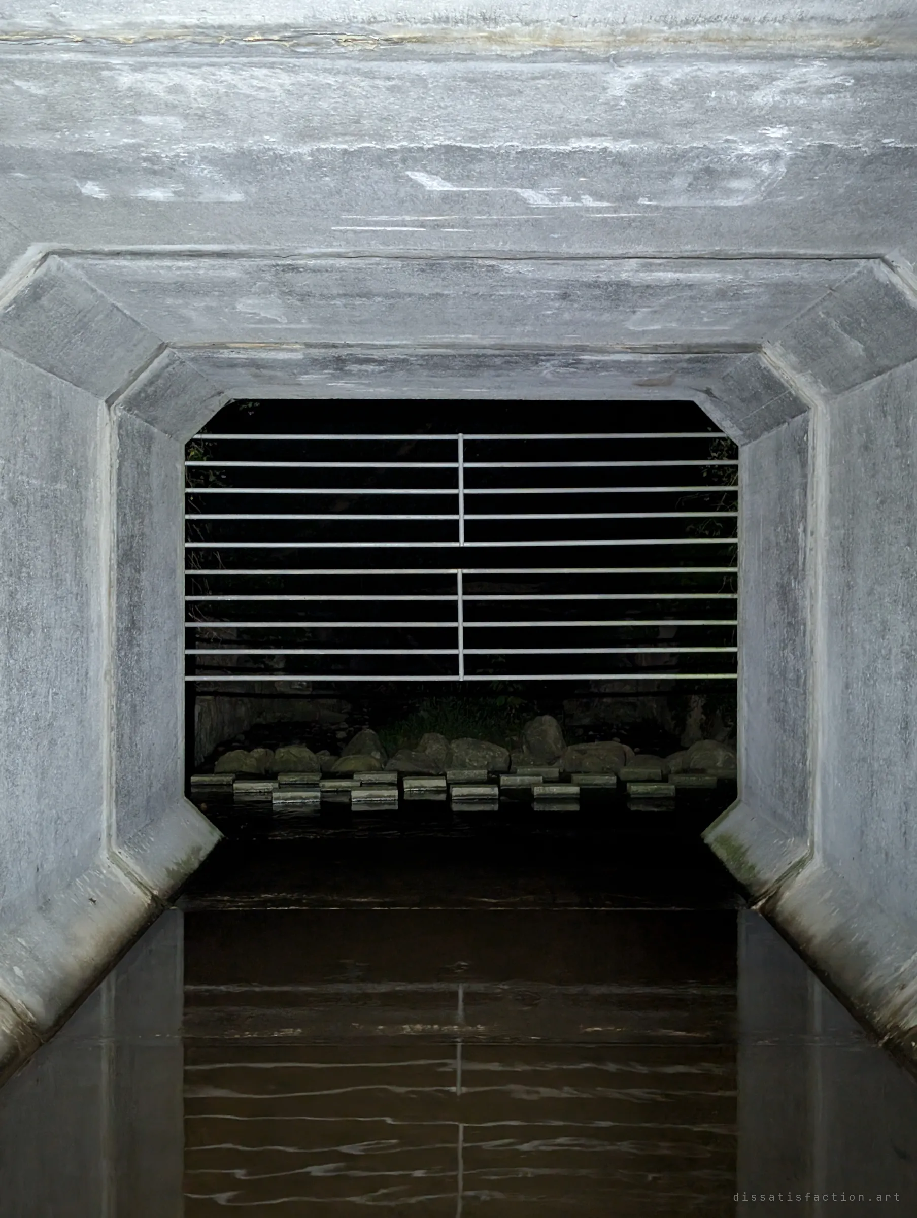 Looking back out of the box culvert through the bar grate at the outfall's rocks and stepping stones, with dark water reflecting the bars. The last glimpse of outside before the tunnel.