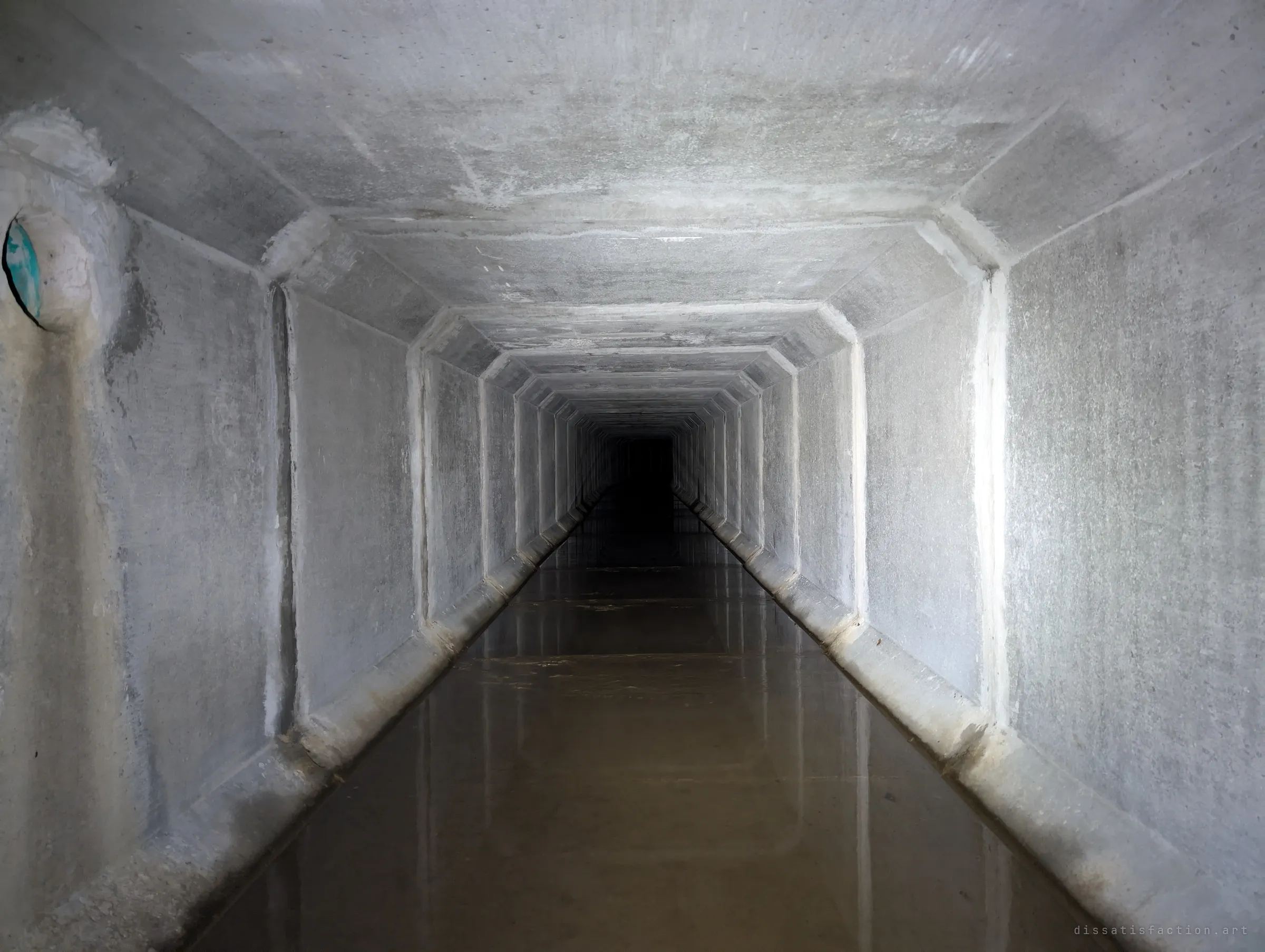 A long, straight precast box drain with chamfered joints receding into darkness over a sheet of shallow brown water. The repeating segments show how far the tunnel runs.