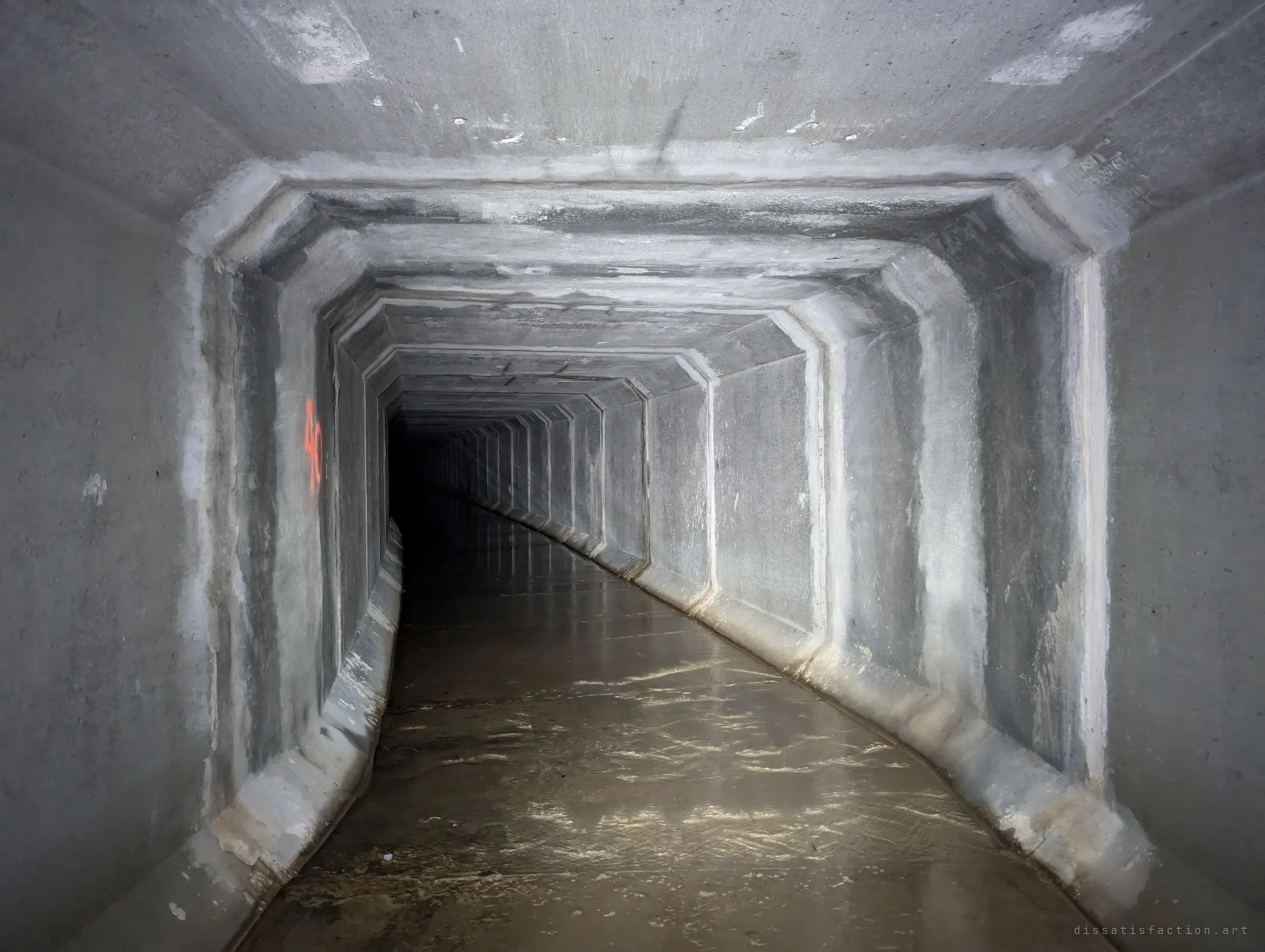 The concrete box drain bends gently right, water rippling across its floor and a spray-painted survey number on one joint. The curve hides what lies ahead.