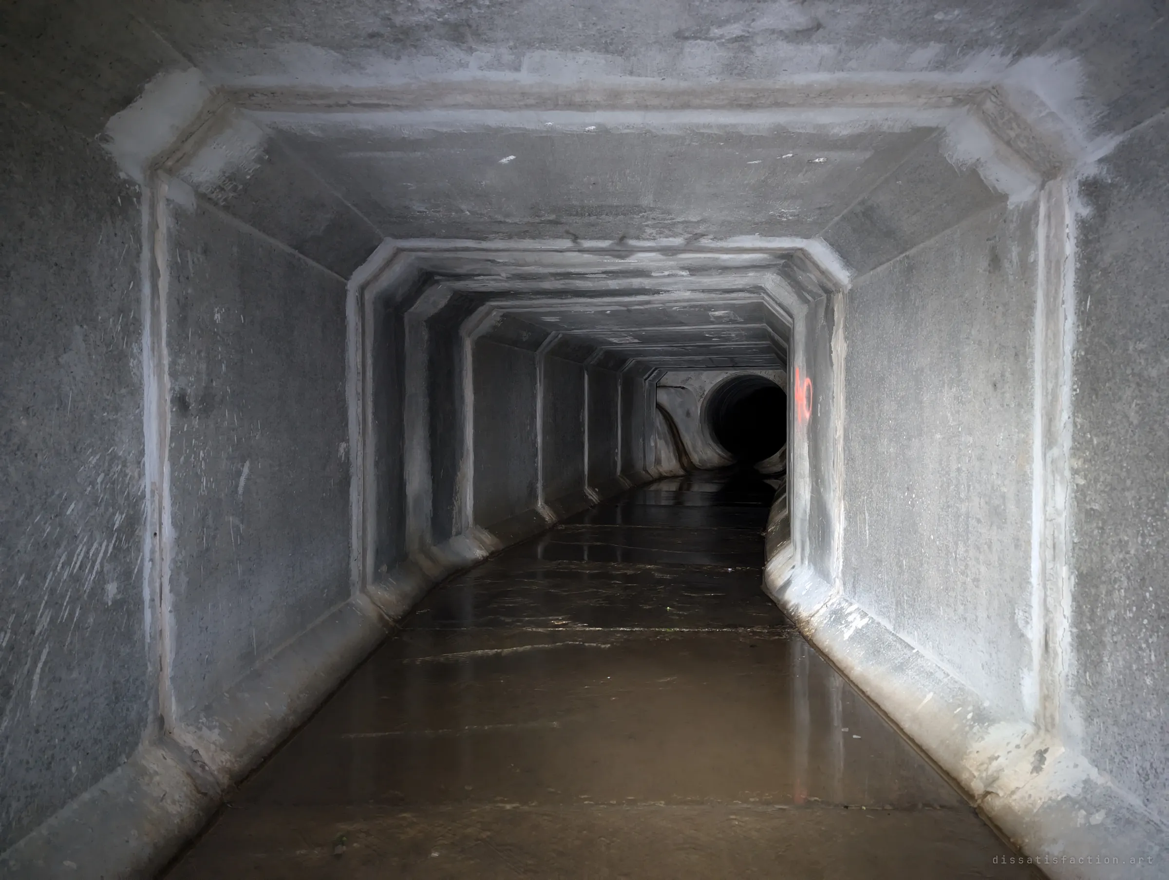 The box drain curves toward a round concrete pipe opening at its far end, joints lit grey by flashlight above wet floor. The shape change marks the transition deeper into the system.