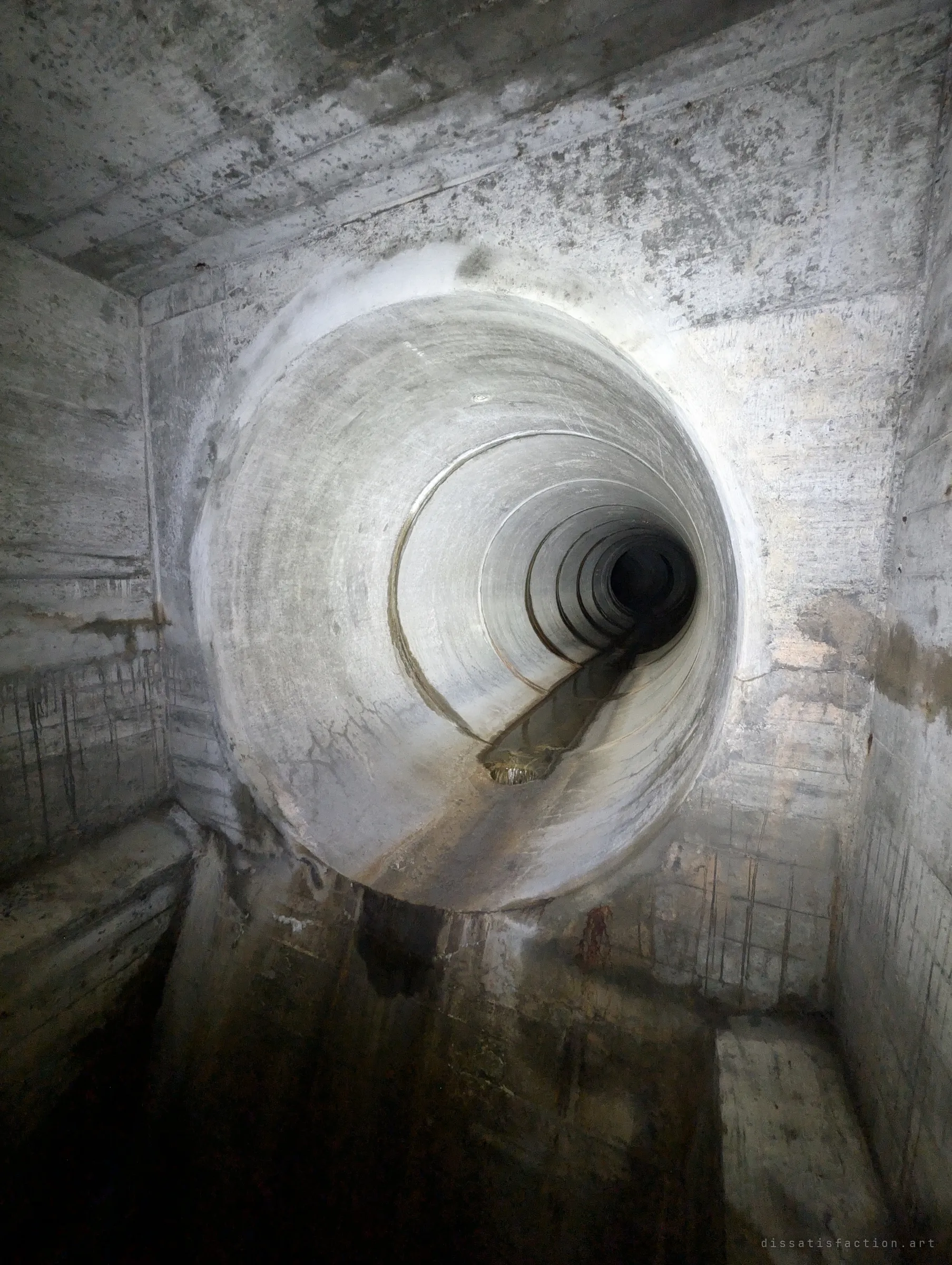 A round pipe opens high in the wall of a stained concrete chamber, above a sheer drop with no railings. The long ringed pipe beyond is the only way on.
