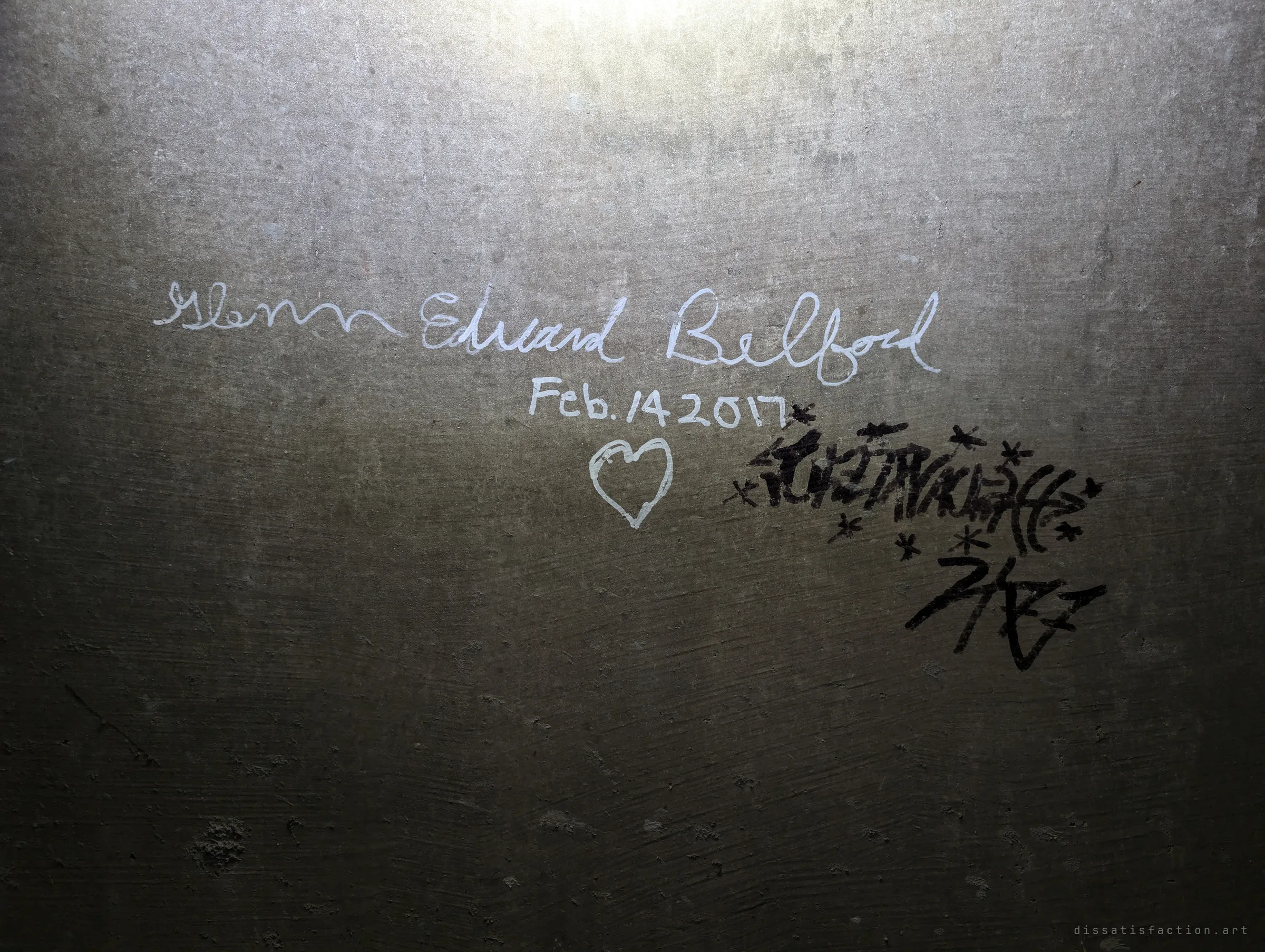 Handwritten white marker lettering with a date and a heart beside a black graffiti tag on the drain's concrete wall. One of the few marks anyone left down here.