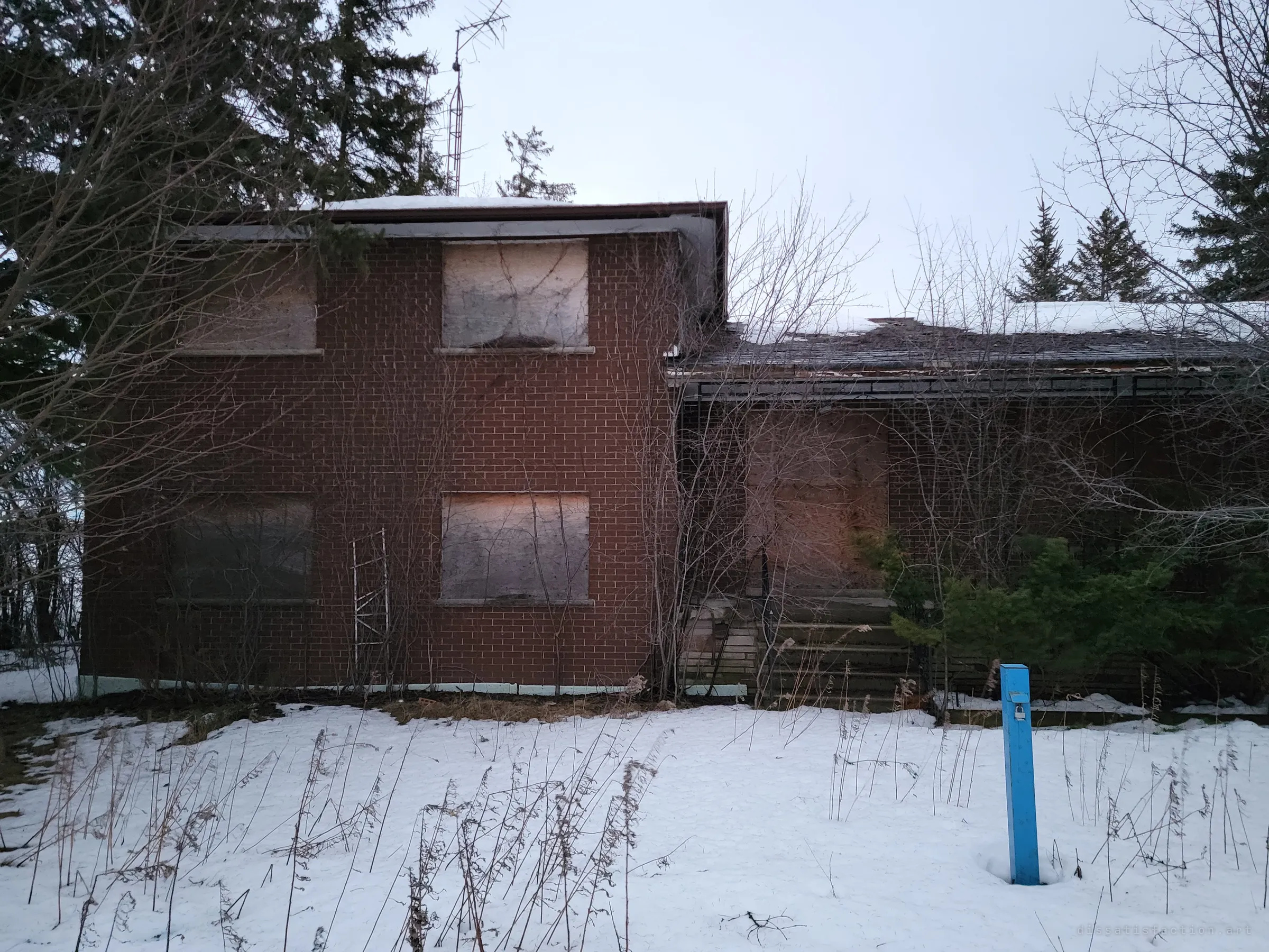 A boarded-up two-storey brick house sits behind snow and bare shrubs, with a blue locked utility post in the yard. Every window is covered in plywood, setting up the abandonment.