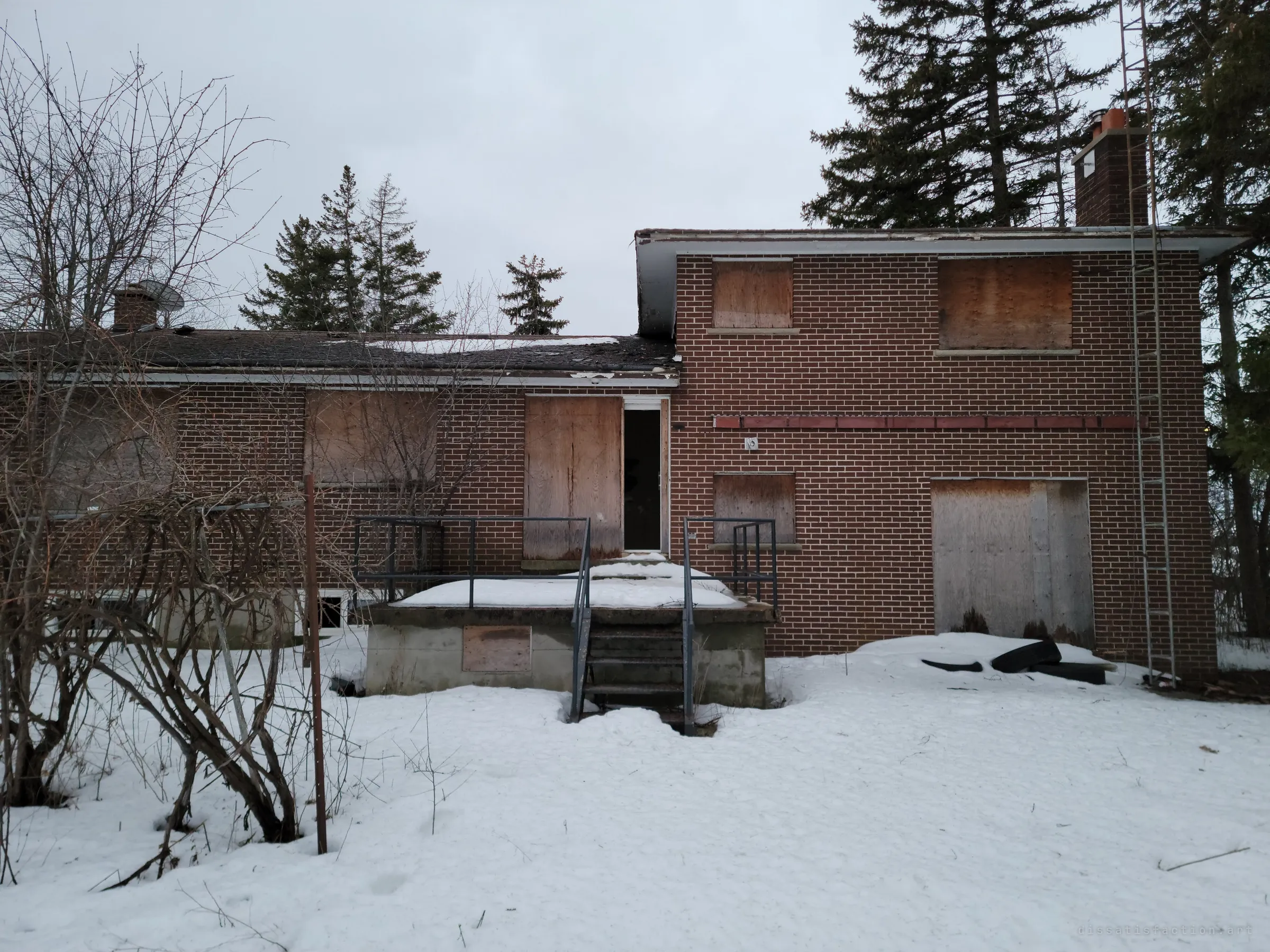 The back of the house under snow: a concrete porch with metal railings leads to an open doorway beside plywood-covered windows. The one open door is the way in.