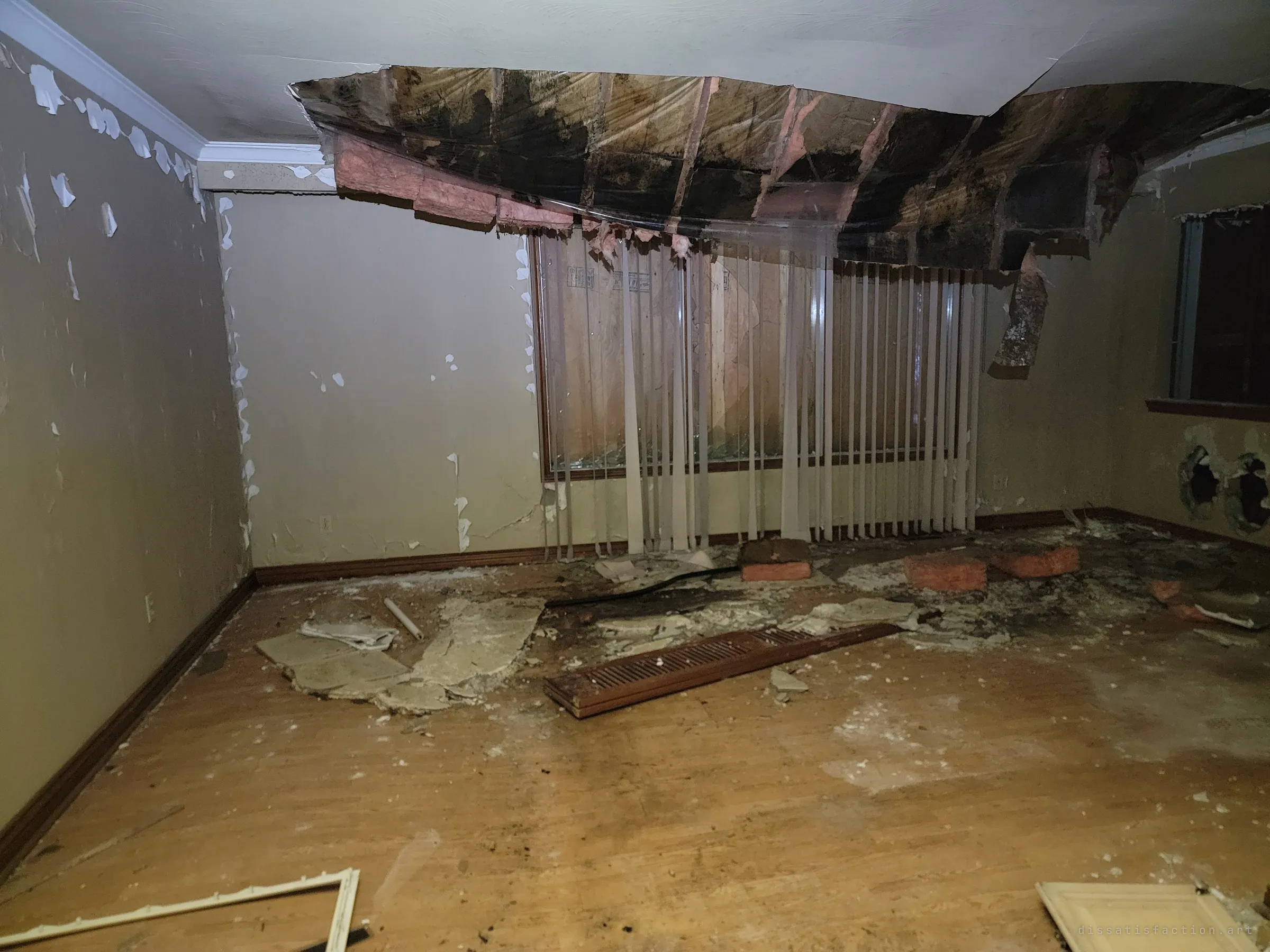 A living room whose ceiling has caved in, with mouldy insulation sagging over vertical blinds and a boarded window. Plaster and a torn-out floor vent lie across the wood floor.