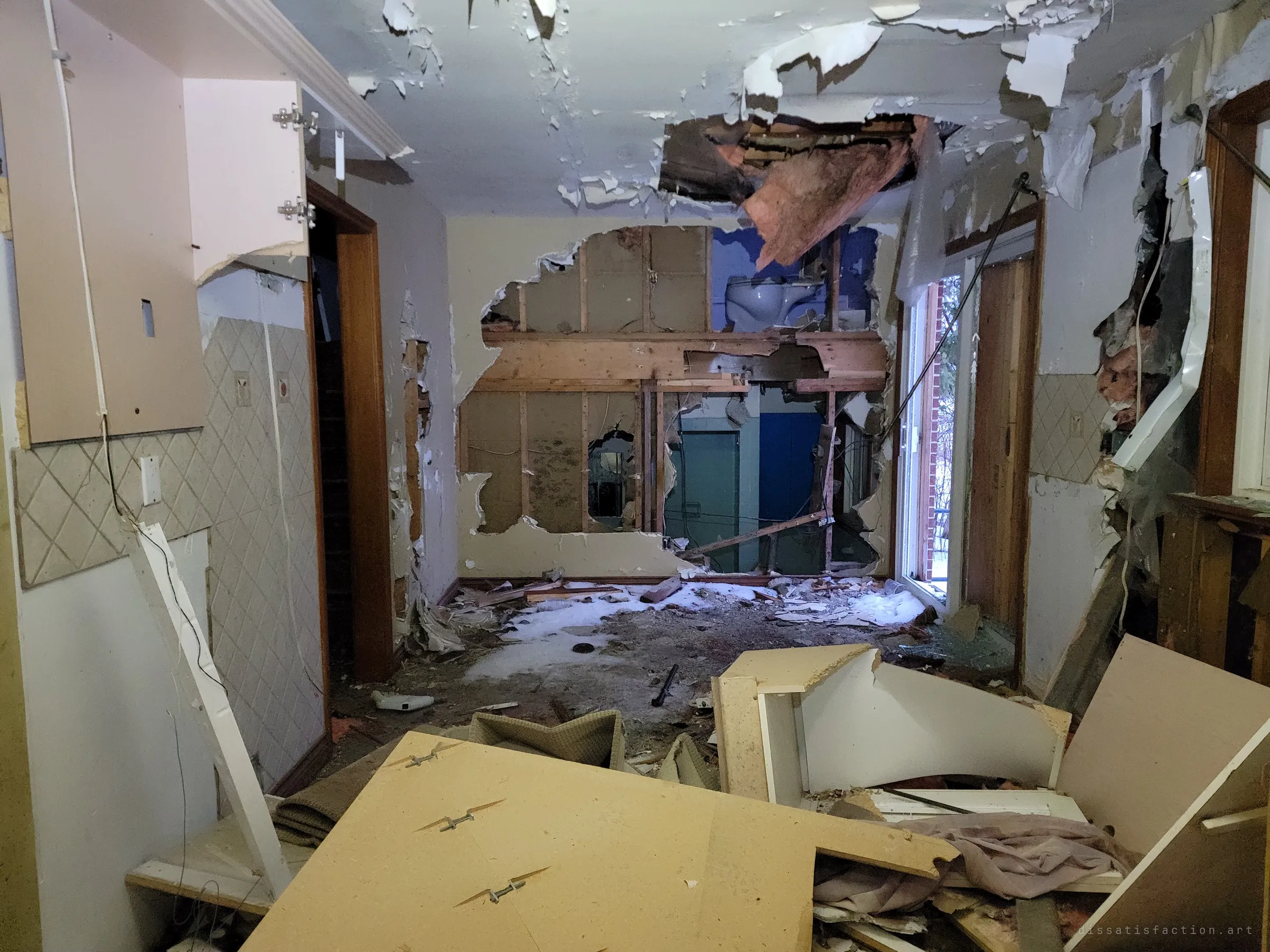 Looking through a kitchen where a wall has been smashed open to the studs, exposing a blue bathroom and toilet beyond. Snow has blown in across the floor through an open patio door.
