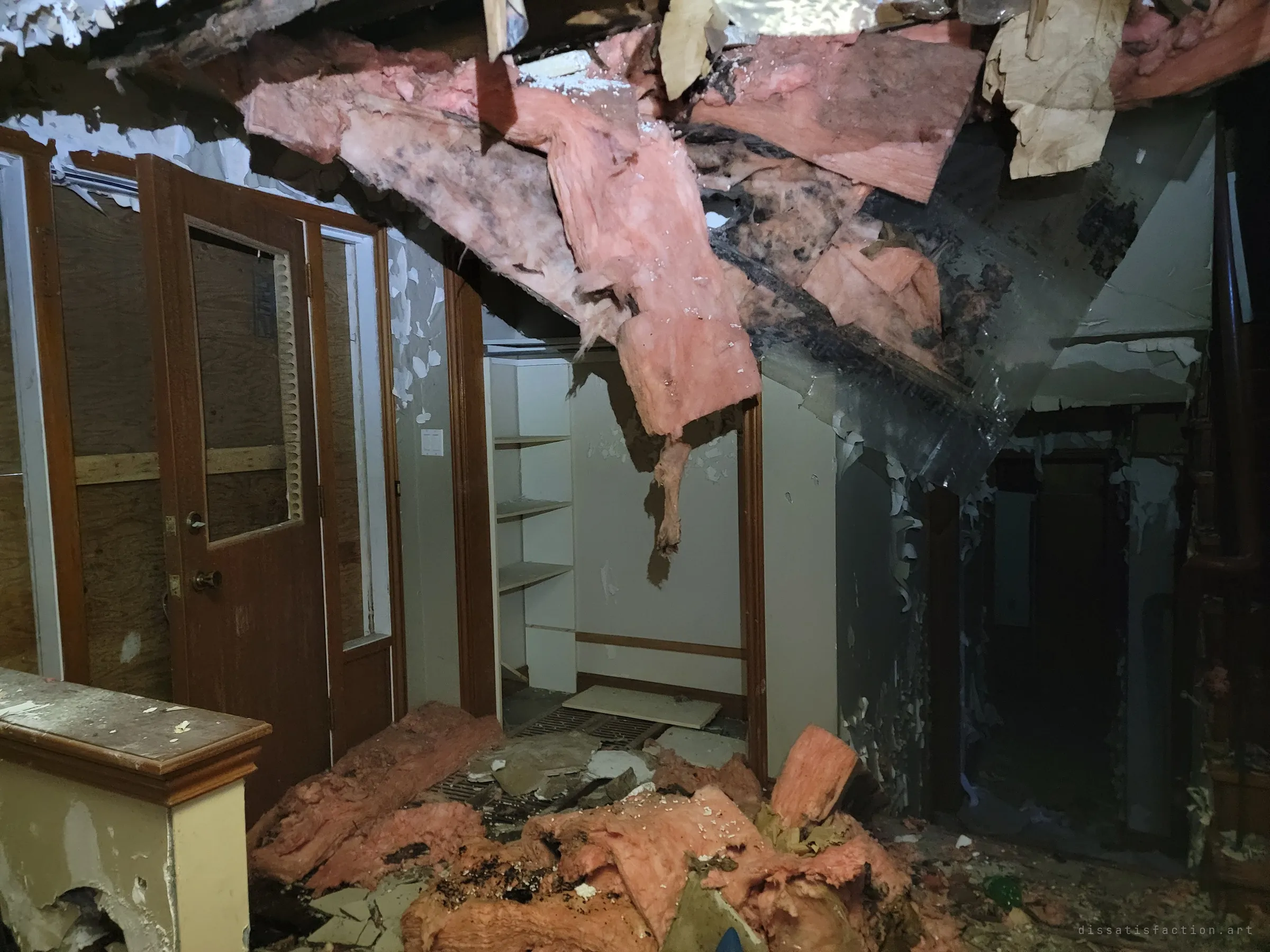 Pink insulation and ceiling boards hang down over a landing beside a glass-panelled door and an empty closet. The collapse shows how far the damage has gone.