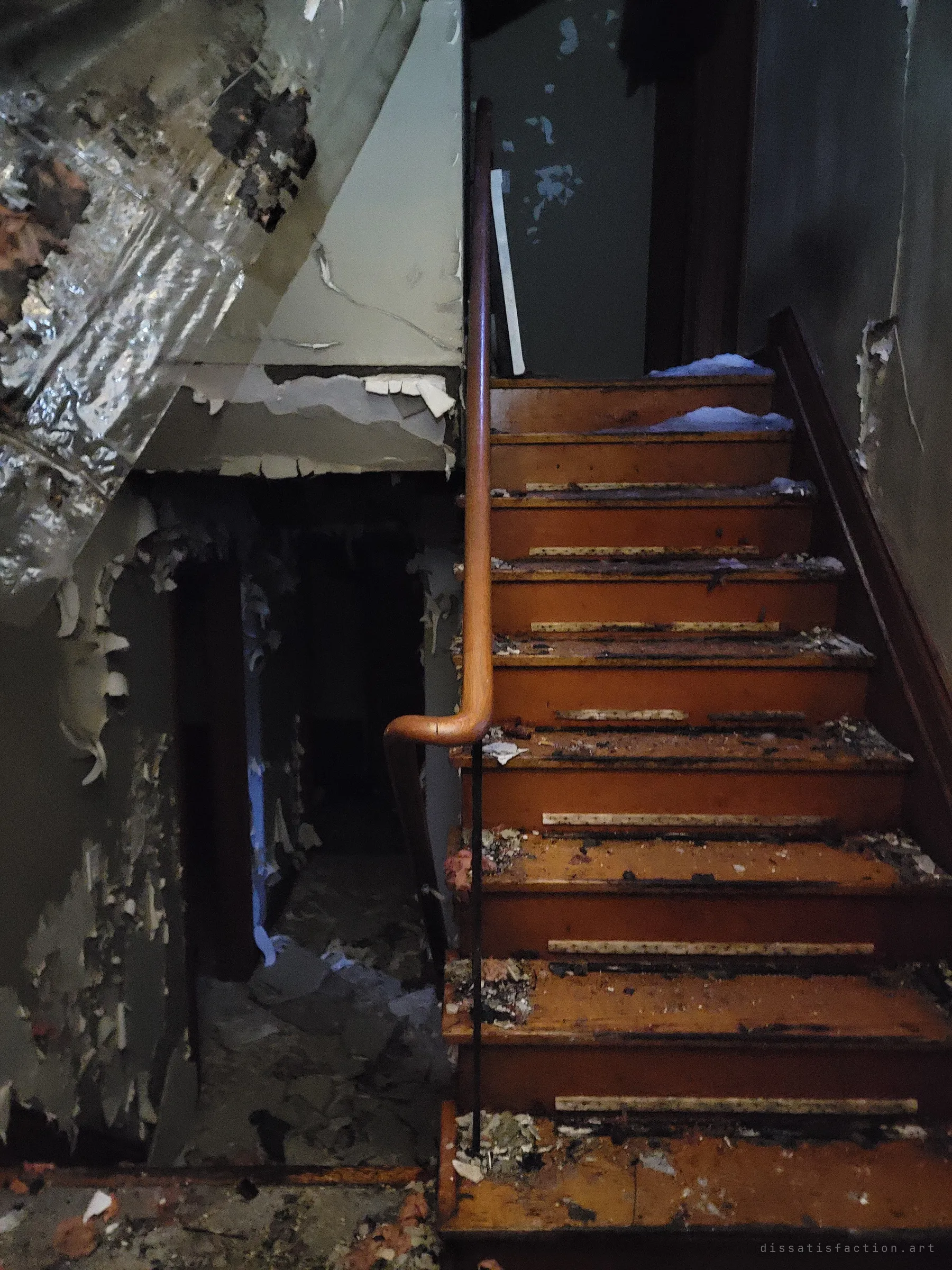 A wooden staircase with a curved handrail climbs past peeling walls and hanging foil vapour barrier, with snow drifted onto the upper steps. It shows the weather getting inside.