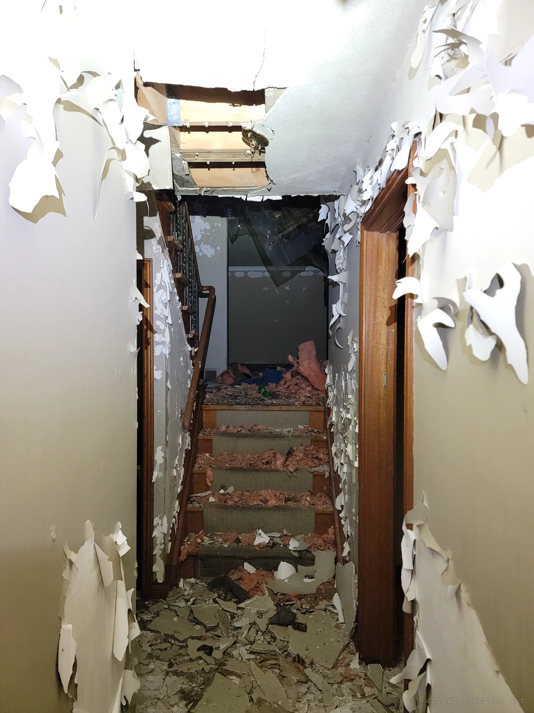 A narrow hallway of curling paint leads to a staircase buried in insulation and ceiling debris, under a hole torn through the ceiling. The corridor frames the wreckage like a tunnel.