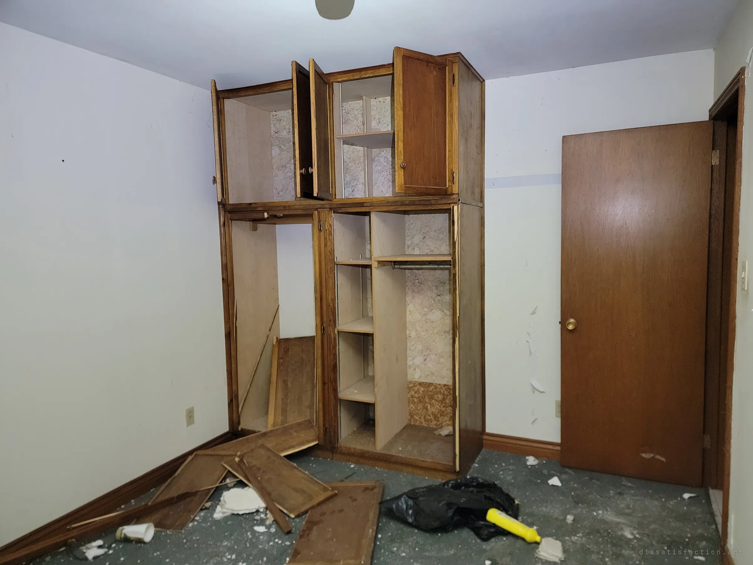 A bedroom with a built-in wooden wardrobe hanging open, its doors torn off and scattered on the floor with a trash bag and debris. The rest of the room is oddly intact.