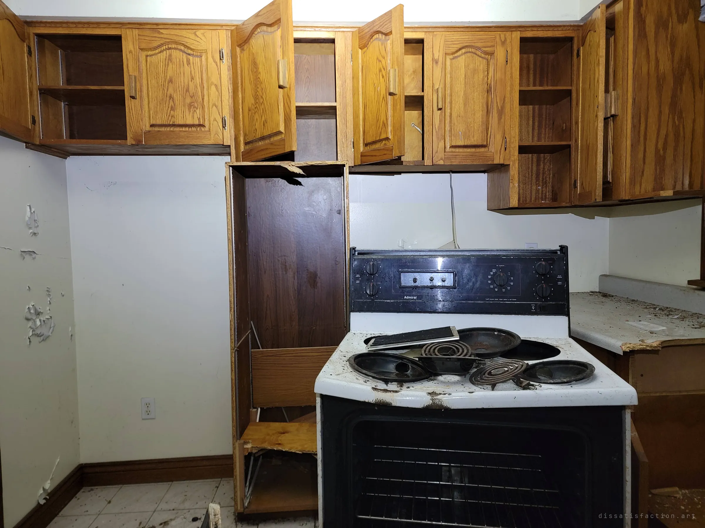 A kitchen with oak cabinets flung open and an old electric stove, its burners and drip pans piled on top. A broken cabinet unit leans beside it.