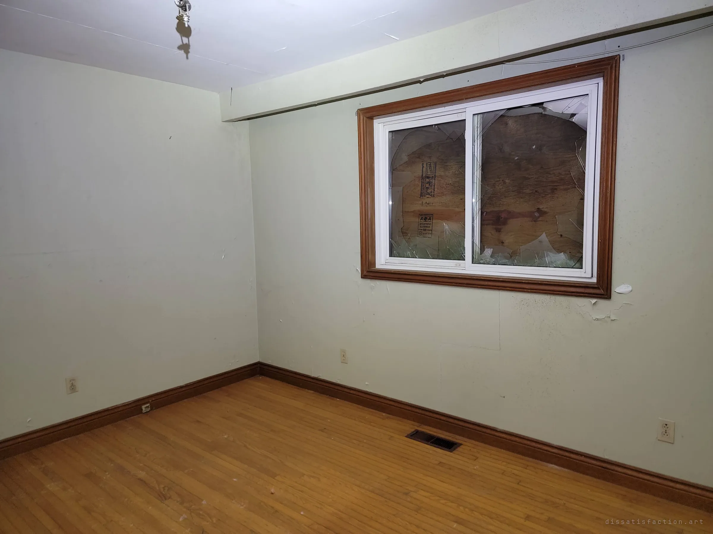 An empty bedroom with bare hardwood floor and pale green walls; the window glass is smashed and boarded from outside with plywood. The quiet emptiness contrasts with the wreckage elsewhere in the house.