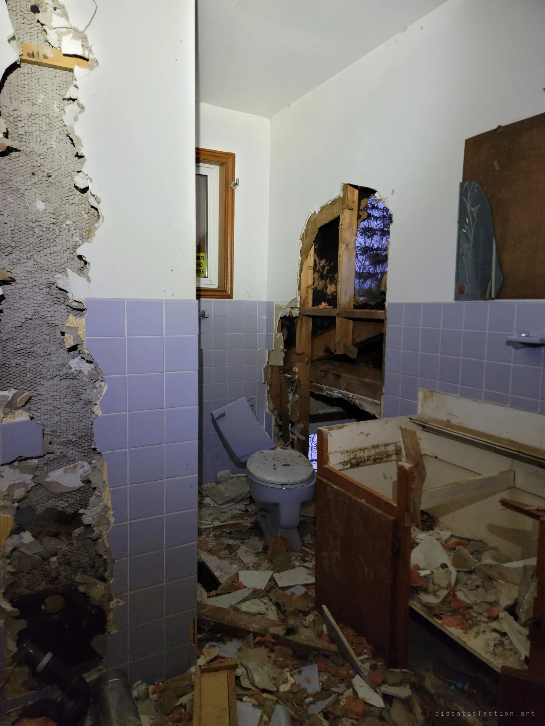 A lavender-tiled bathroom torn apart: the toilet tank is knocked askew, the vanity is smashed and a hole through the wall opens onto the dusk outside. Debris and insulation cover the floor.