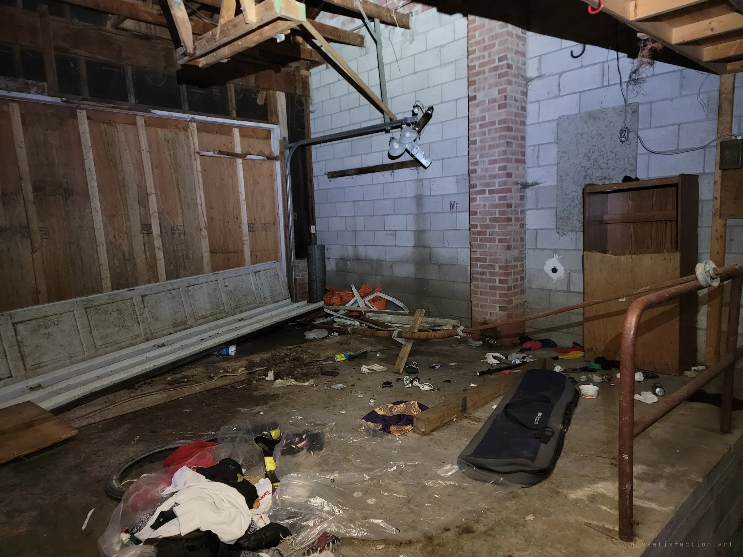 The attached garage with its door panels fallen in, a brick column against cinder-block walls and clothes, cans and a board bag strewn across the concrete. A railing guards the stairwell down to the basement.