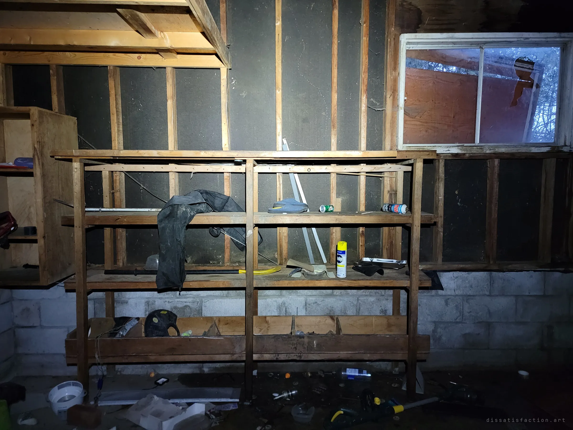 Wooden garage shelving under a flashlight, holding a rubber coat, spray cans and leftover tools against bare studs. A broken window above shows only plywood and branches outside.