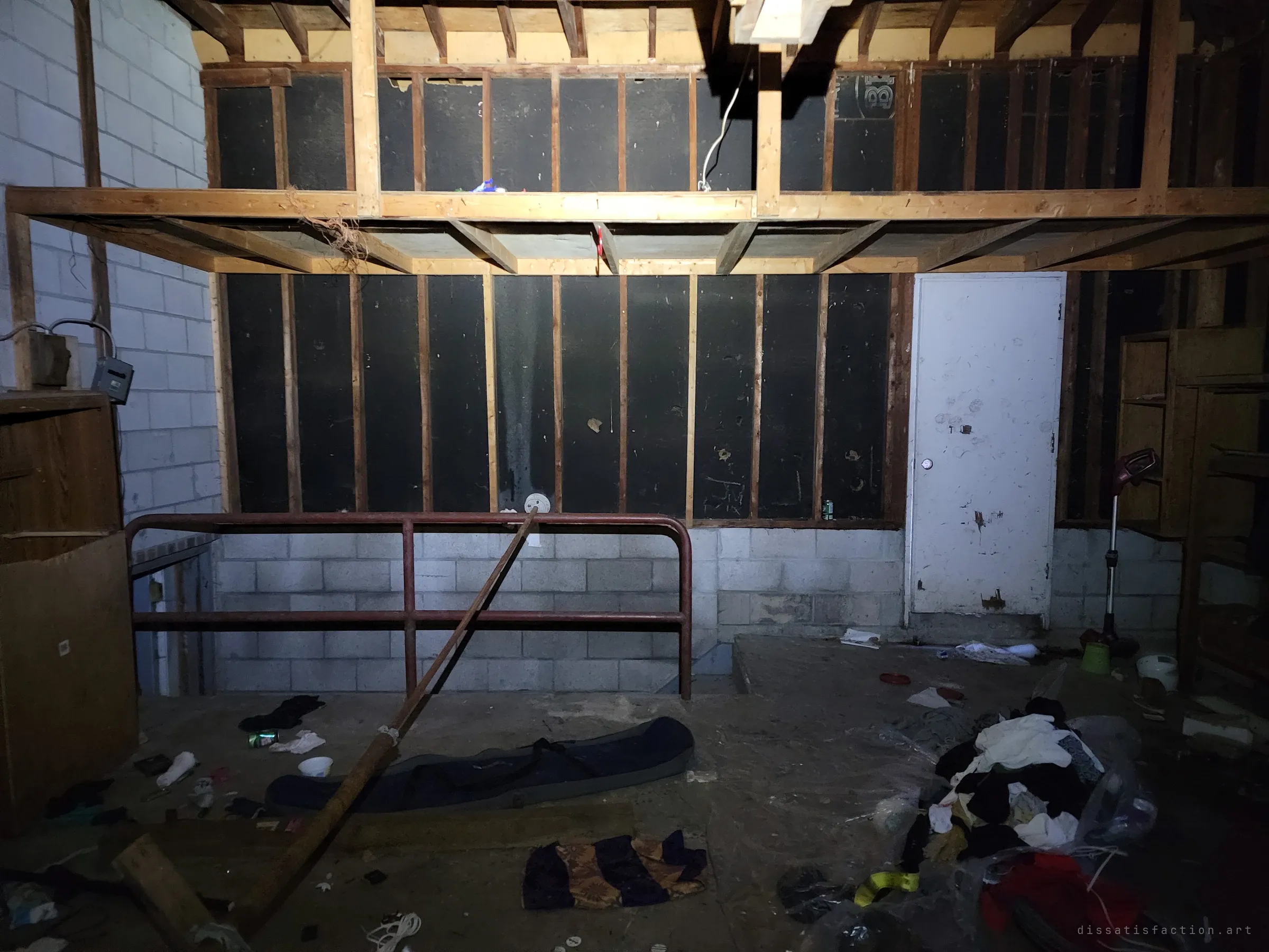 The back wall of the garage: an open-stud loft over black sheathing, a white side door and a red railing around the basement stairwell. A pole leans across the railing above a litter of clothes on the floor.