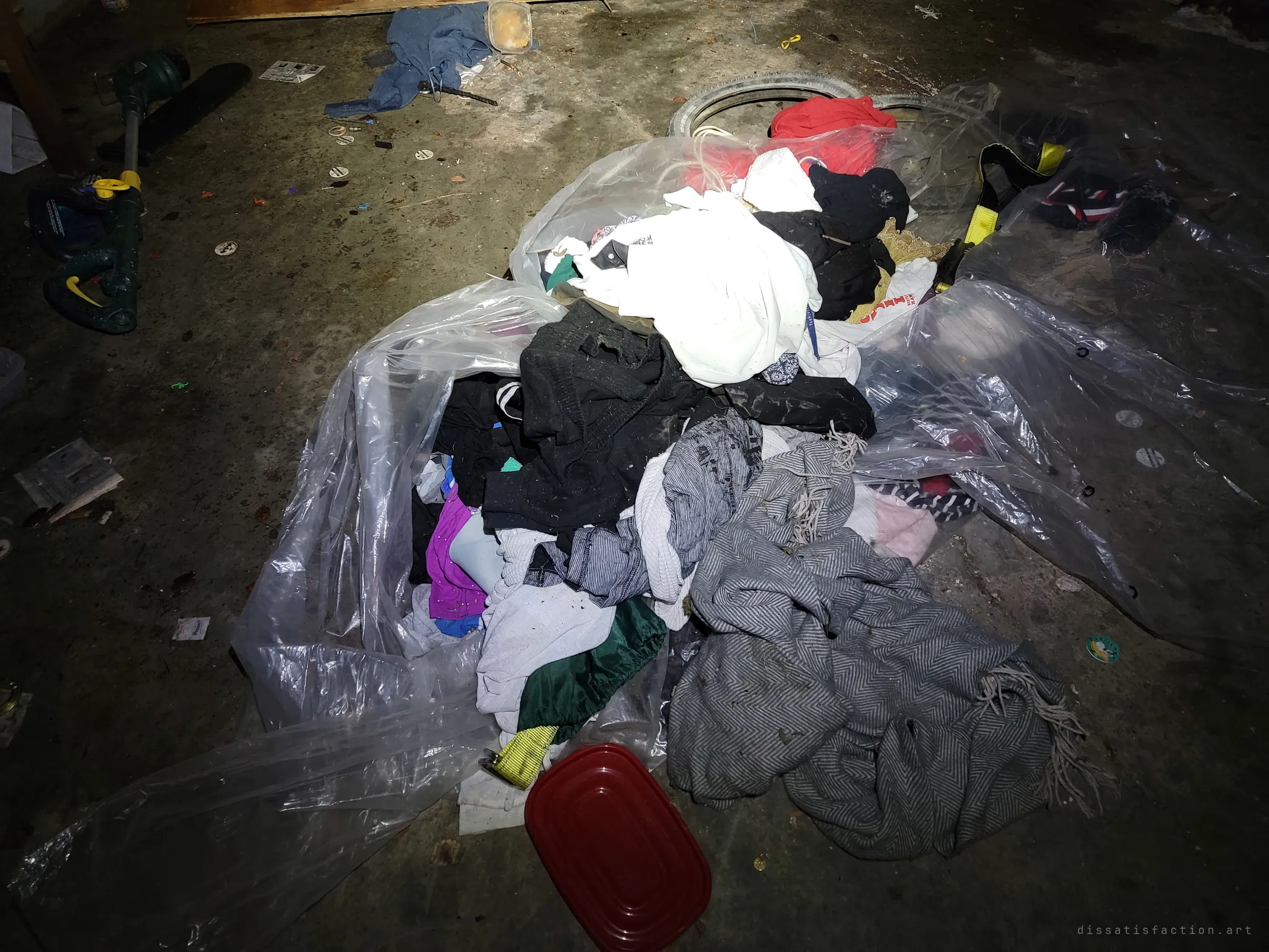 A heap of clothes and blankets spilling out of torn plastic sheeting on the garage floor, beside a bike tyre and a string trimmer. Signs of people passing through after the house was left.